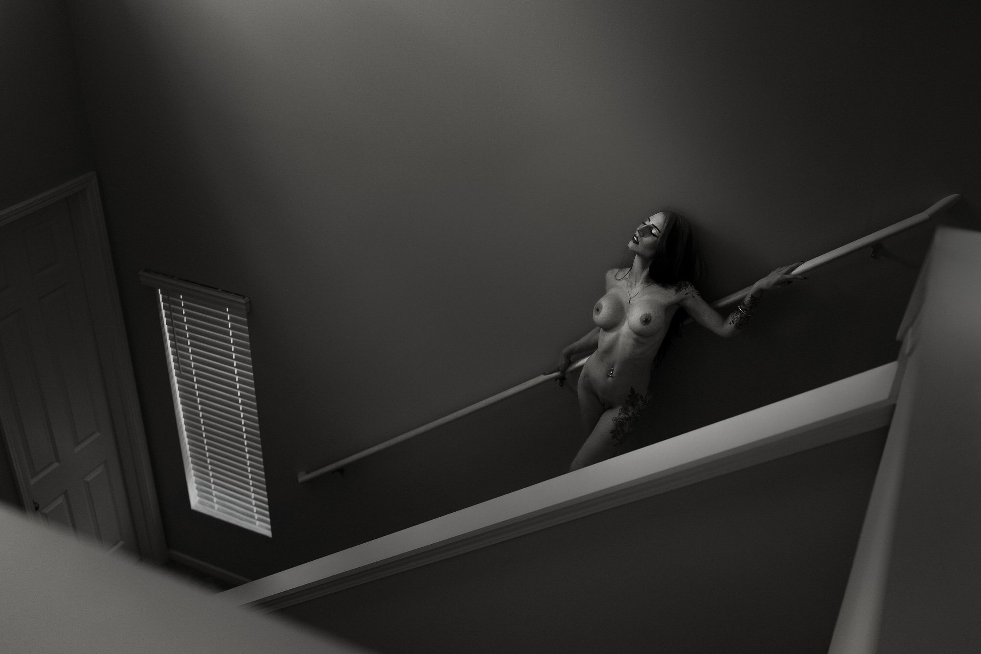 A black and white photograph of a topless woman with tattoos, holding onto a handrail on a staircase, with window blinds partially visible in the background.