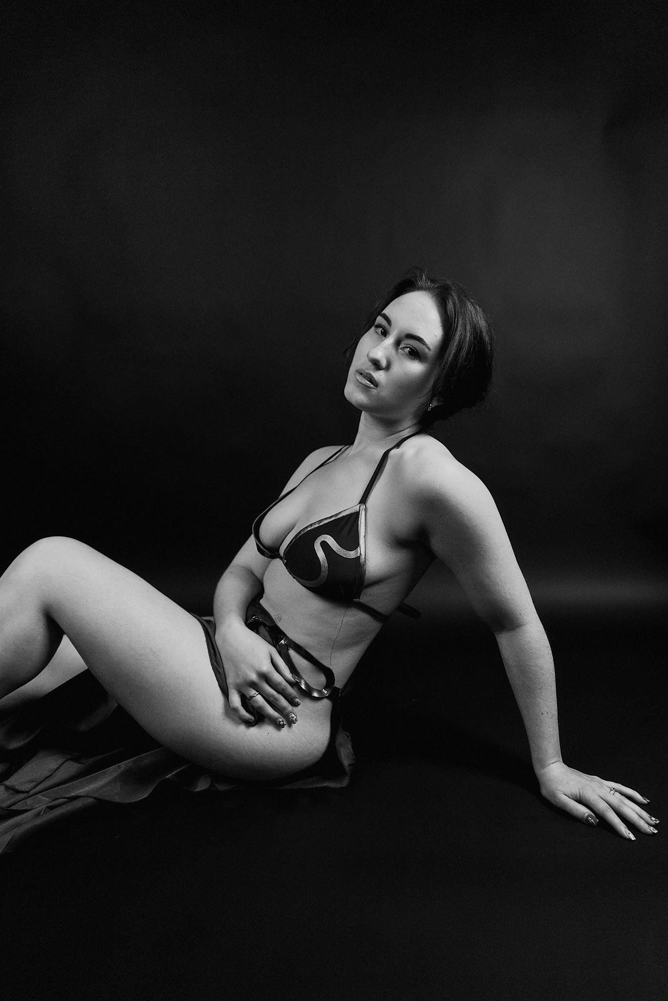 Black and white photo of a woman with short hair, wearing revealing lingerie, posed on a black background. She is sitting with one arm supporting her and the other hand resting on her thigh, looking at the camera with a confident expression.