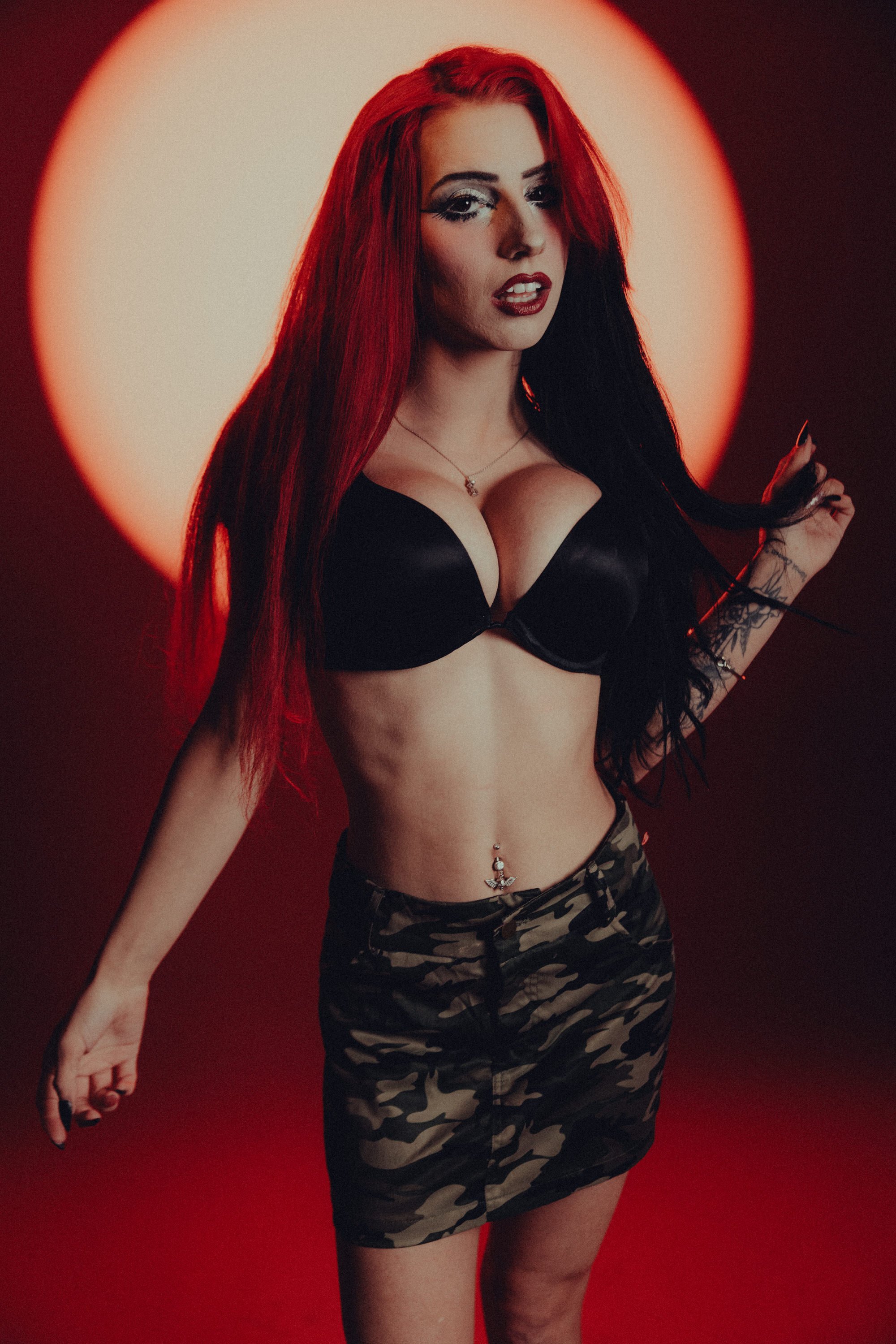 A woman with long red and black hair, wearing a black bra, camouflage shorts, and a butterfly belly chain, standing in front of a red and black background with a circular light behind her.