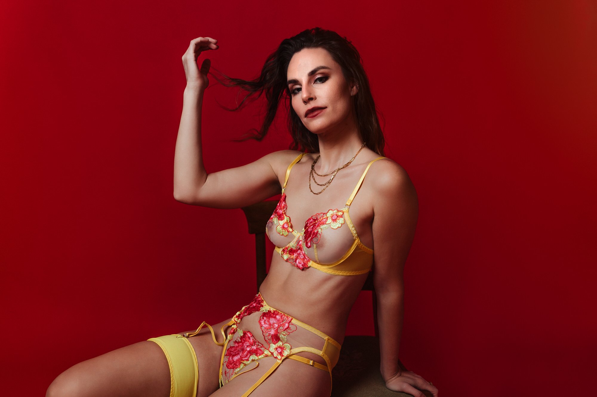 A woman with long dark hair wearing a yellow lingerie set with red floral embroidery, sitting against a red background.