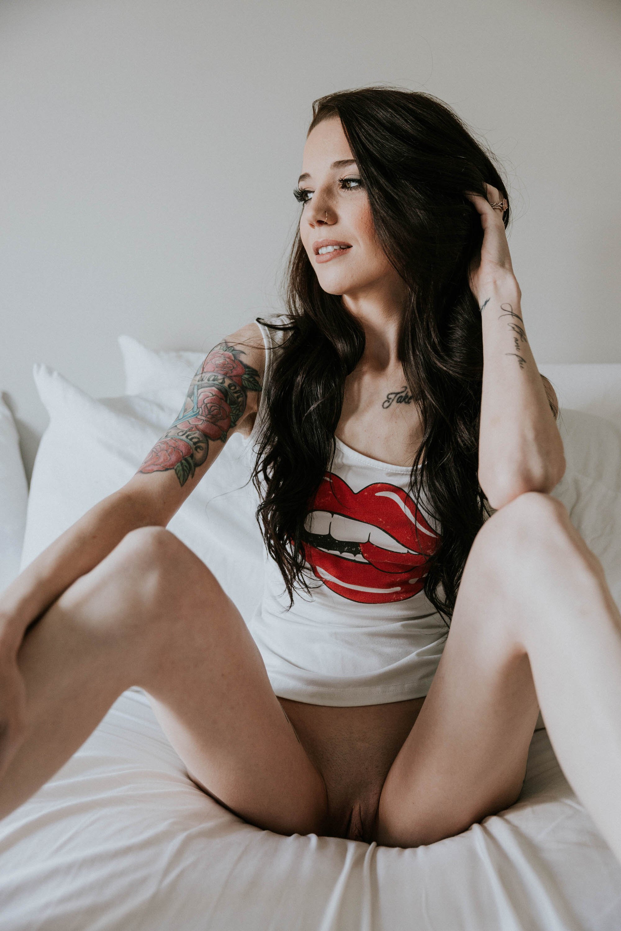 A young woman with dark hair and tattoos on her arm and chest sitting on a bed, wearing a white tank top with a red lips graphic, looking to her left, with a neutral background.