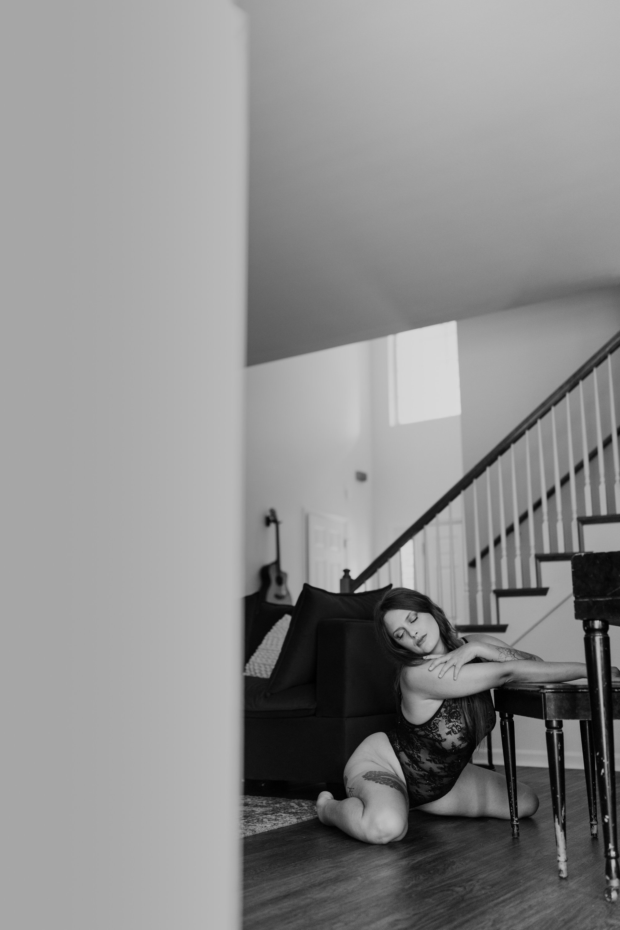 A woman in lace lingerie is posing on the floor beside a table, resting her head on the table's edge, with her eyes closed and a relaxed expression. The setting is a modern, well-lit interior with a staircase, a guitar, and framed artwork visible in 