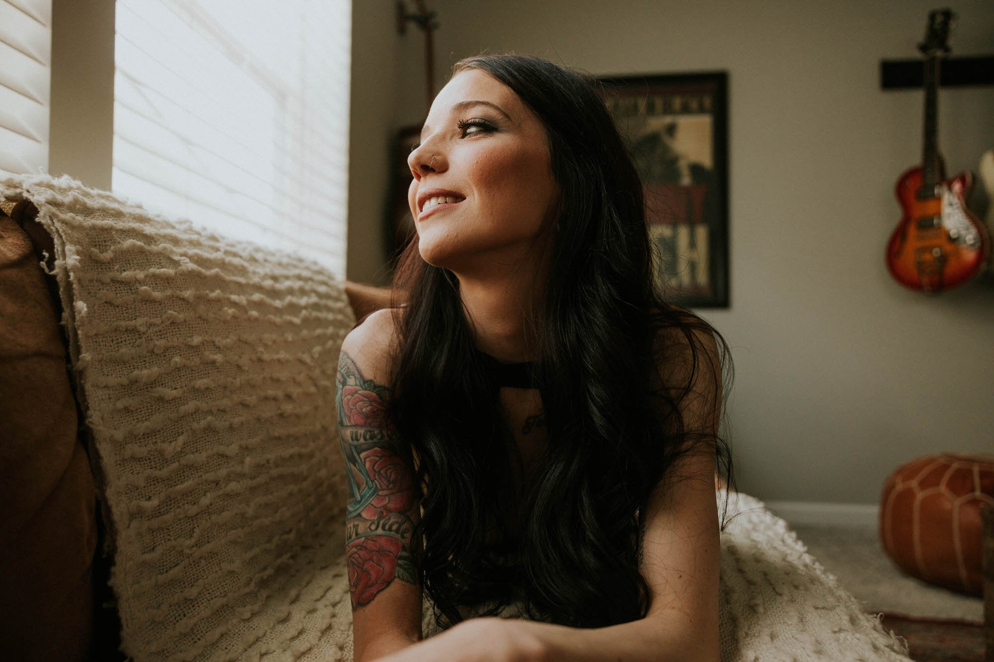 A woman with long dark hair, tattoos on her arm, a nose ring, and makeup, smiling and looking out the window.