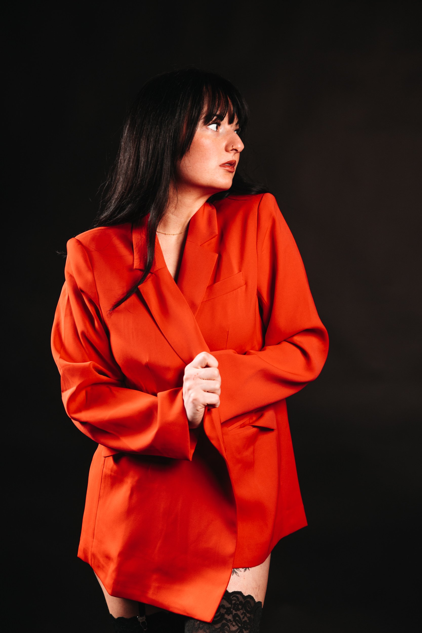 A woman with dark hair wearing a bright red blazer and matching skirt, black lace stockings, and posing against a black background.