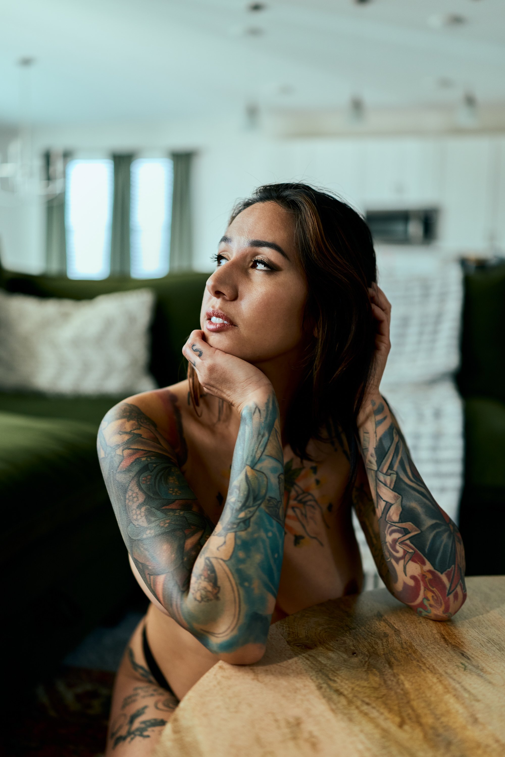 A woman with tattoos on her arms and torso, sitting topless at a table in a bright room, gazing off to the side with her hand resting on her face.