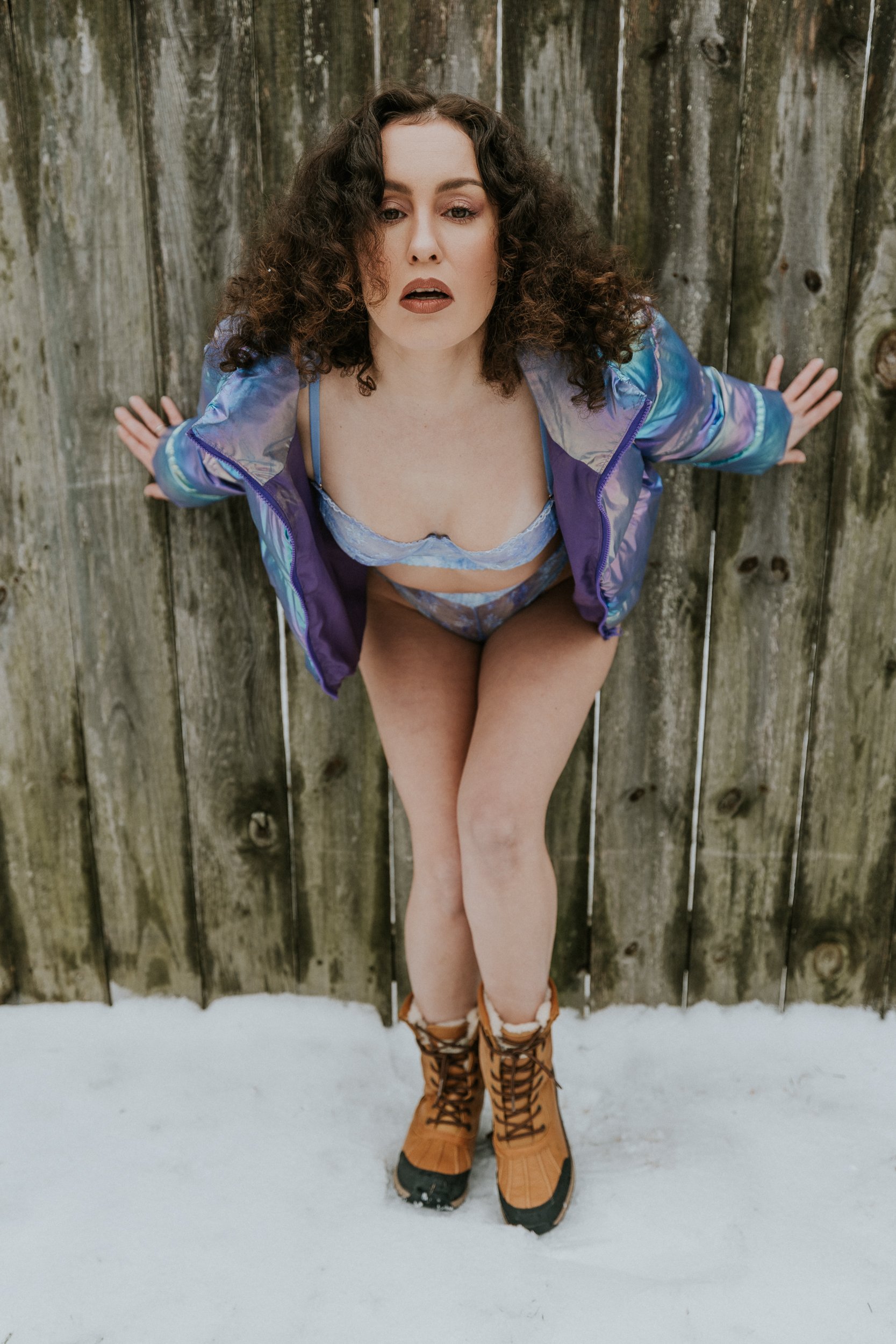 A woman with curly hair stands against a wooden fence in the snow, wearing a purple jacket, lace lingerie, tan boots, and snow boots.