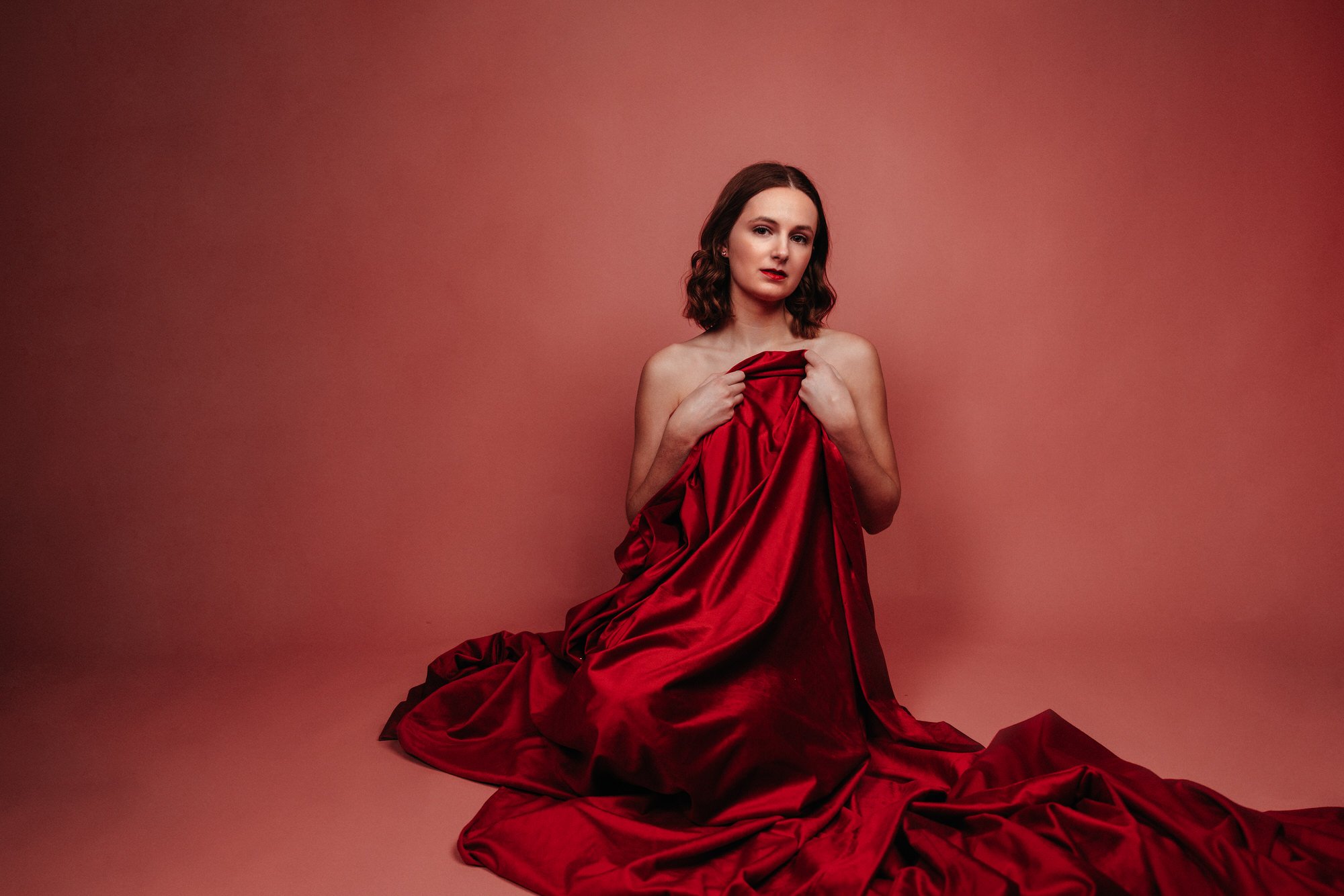 A woman with shoulder-length brown hair and red lipstick is partially covered with red fabric, sitting against a pinkish-red background.