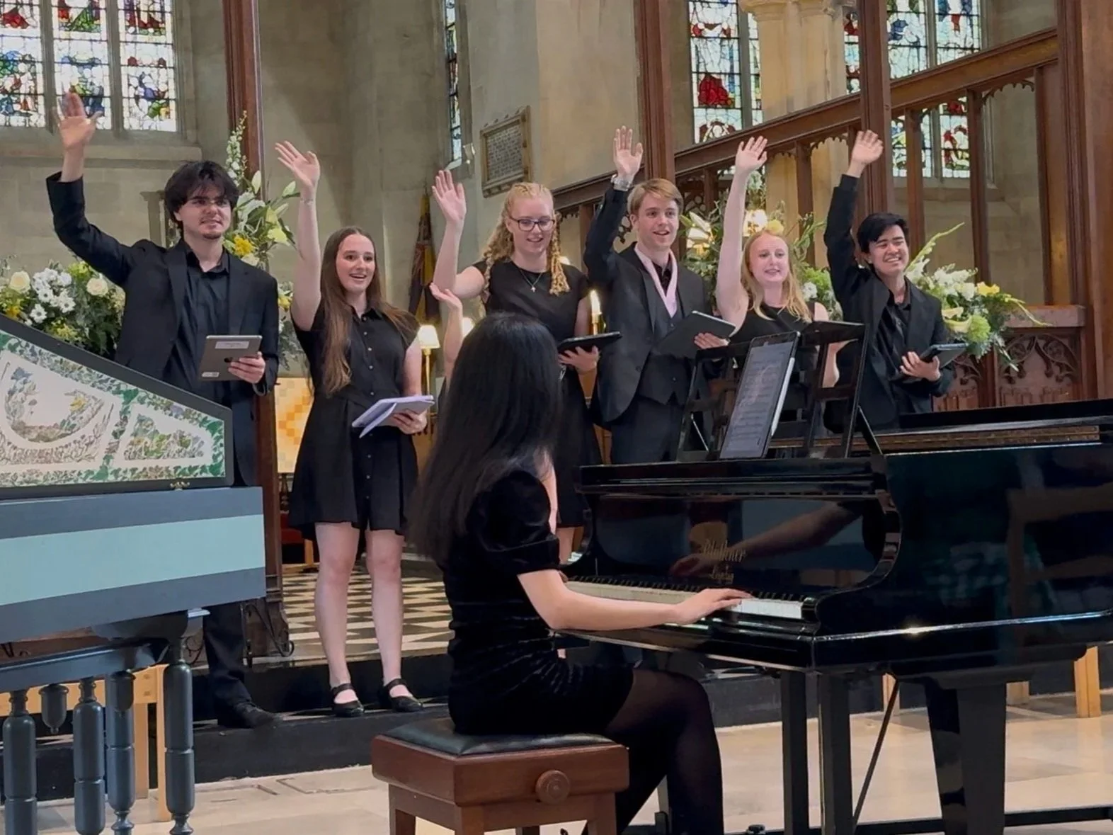 A group of young choral scholars standing on a church altar, raising their hands in song, with a pianist playing in the foreground, inside St Mary Magdalene, with stained glass windows and floral arrangements.