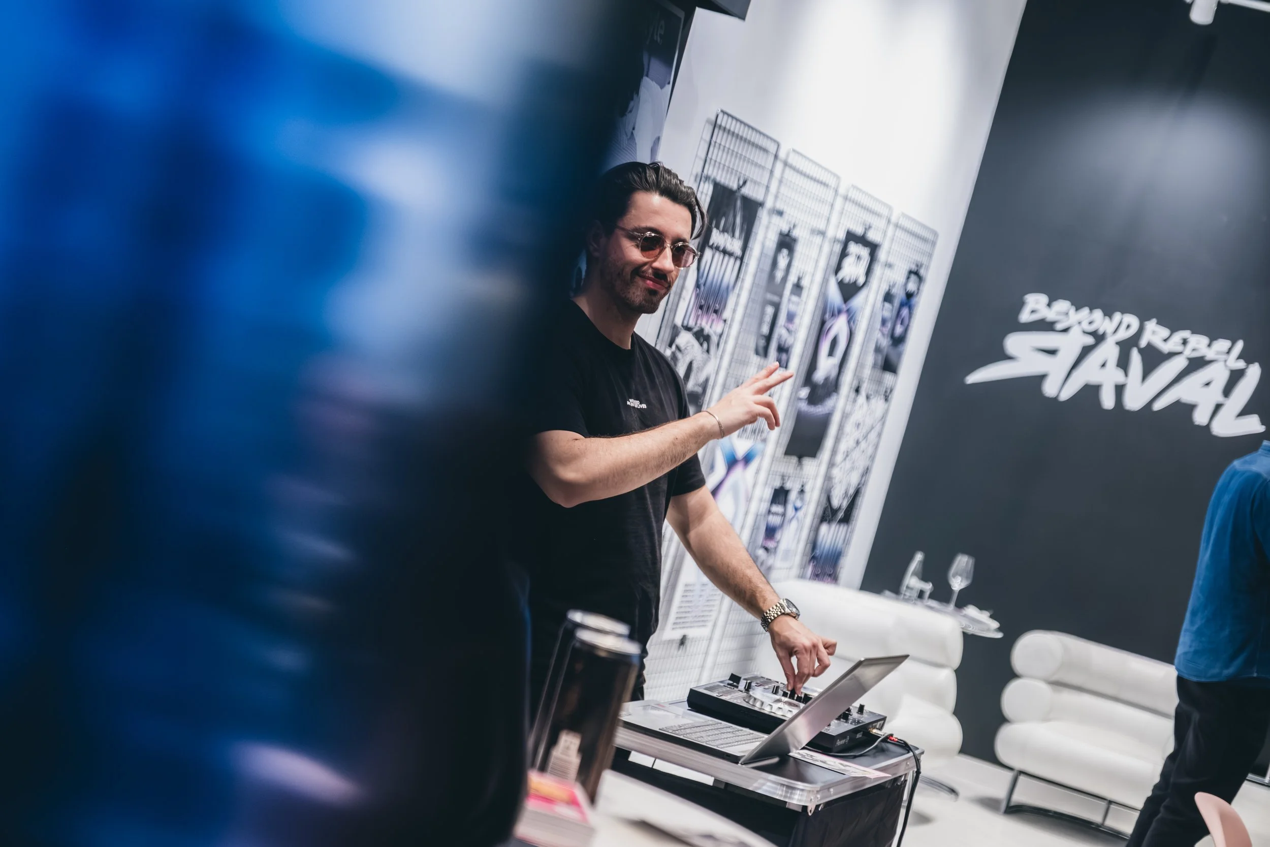Ein Mann mit schwarzen Haaren, Brille und schwarzem T-Shirt, der an einem Tisch mit elektronischem Musikgerät arbeitet, während im Hintergrund Poster mit Text und Grafiken hängen.