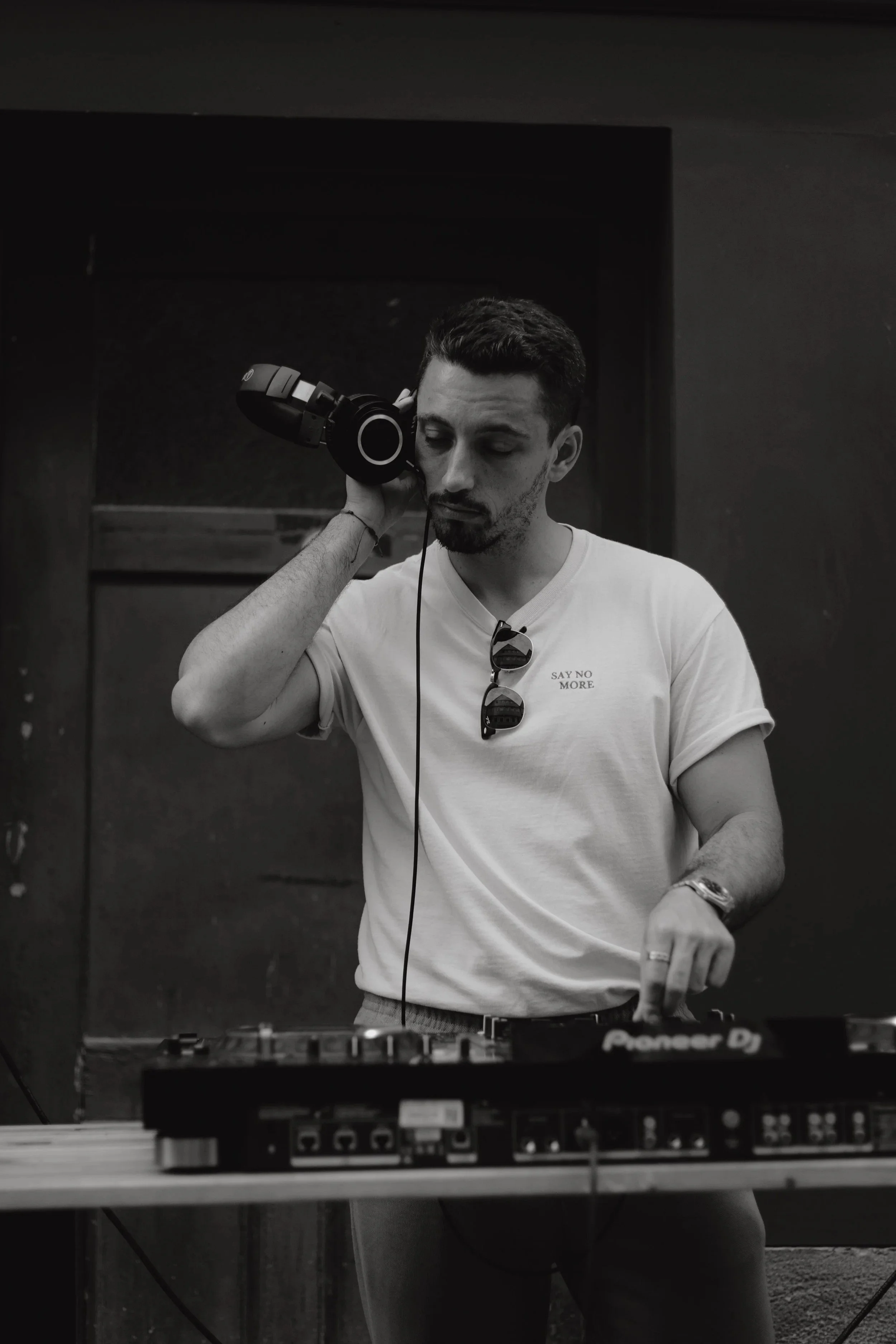 Mann mit dunklen Haaren und Bart trägt Kopfhörer, steht hinter einem DJ-Pult, trägt ein weißes T-Shirt mit der Aufschrift 'SAY NO MORE' und Sonnenbrille. Schwarz-weiß-Foto, dunkler Hintergrund.