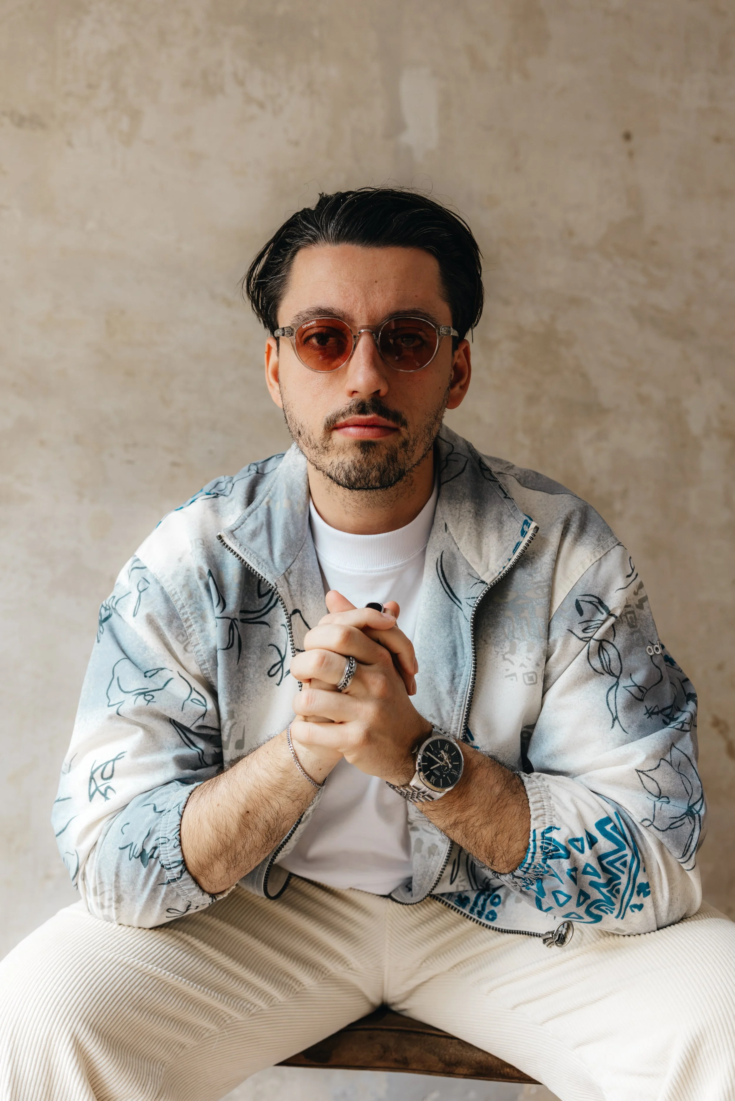 Ein junger Mann mit dunklen, glatten Haaren trägt eine zerrissene, grafisch bedruckte Jacke, Sonnenbrille mit roten Gläsern, eine Armbanduhr und mehrere Schmuckstücke. Er sitzt auf einem Stuhl vor einem neutralen, texturierten Hintergrund.