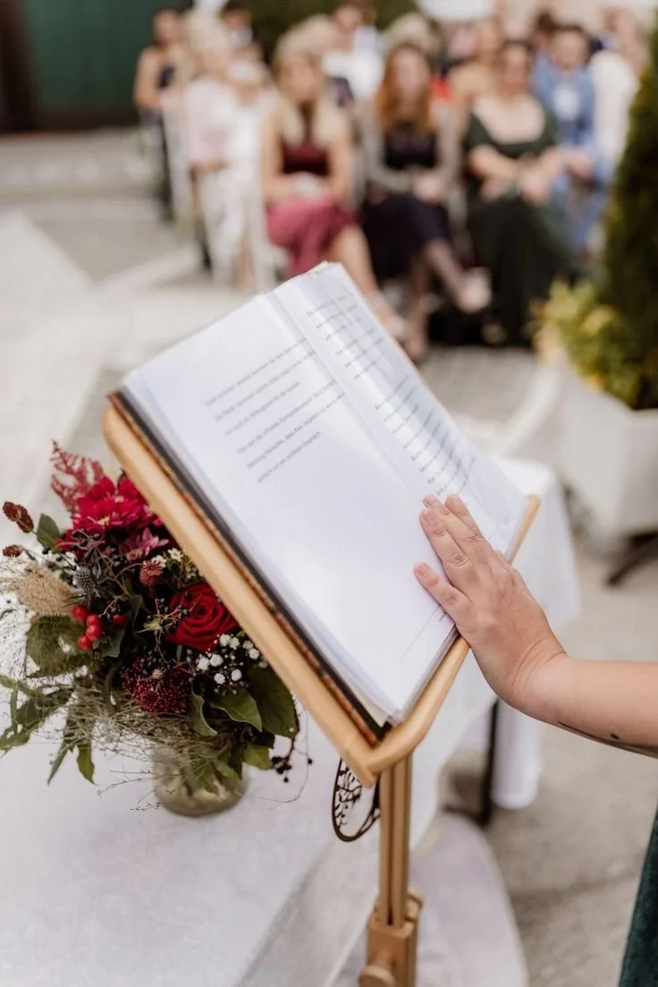 Eine Person liest eine offene Buchseite bei einer Zeremonie mit einem Blumenarrangement auf einem Podest, im Hintergrund sitzen Gäste.