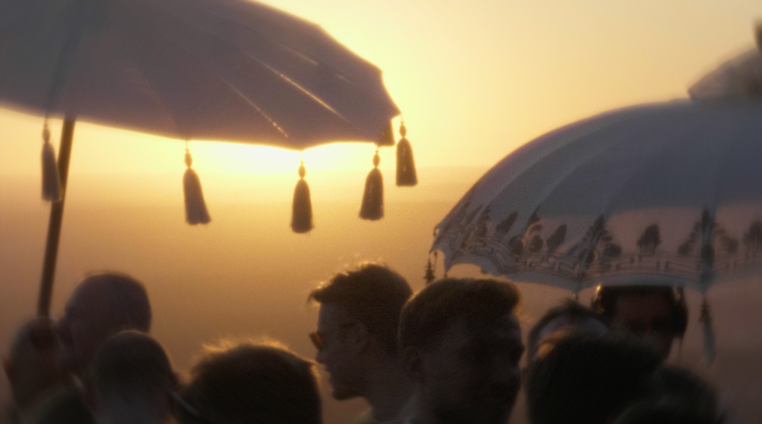 Menschen auf einem Strand bei Sonnenuntergang, mit Sonnenschirmen im Vordergrund und einem großen, gemusterten Gebäude im Hintergrund.