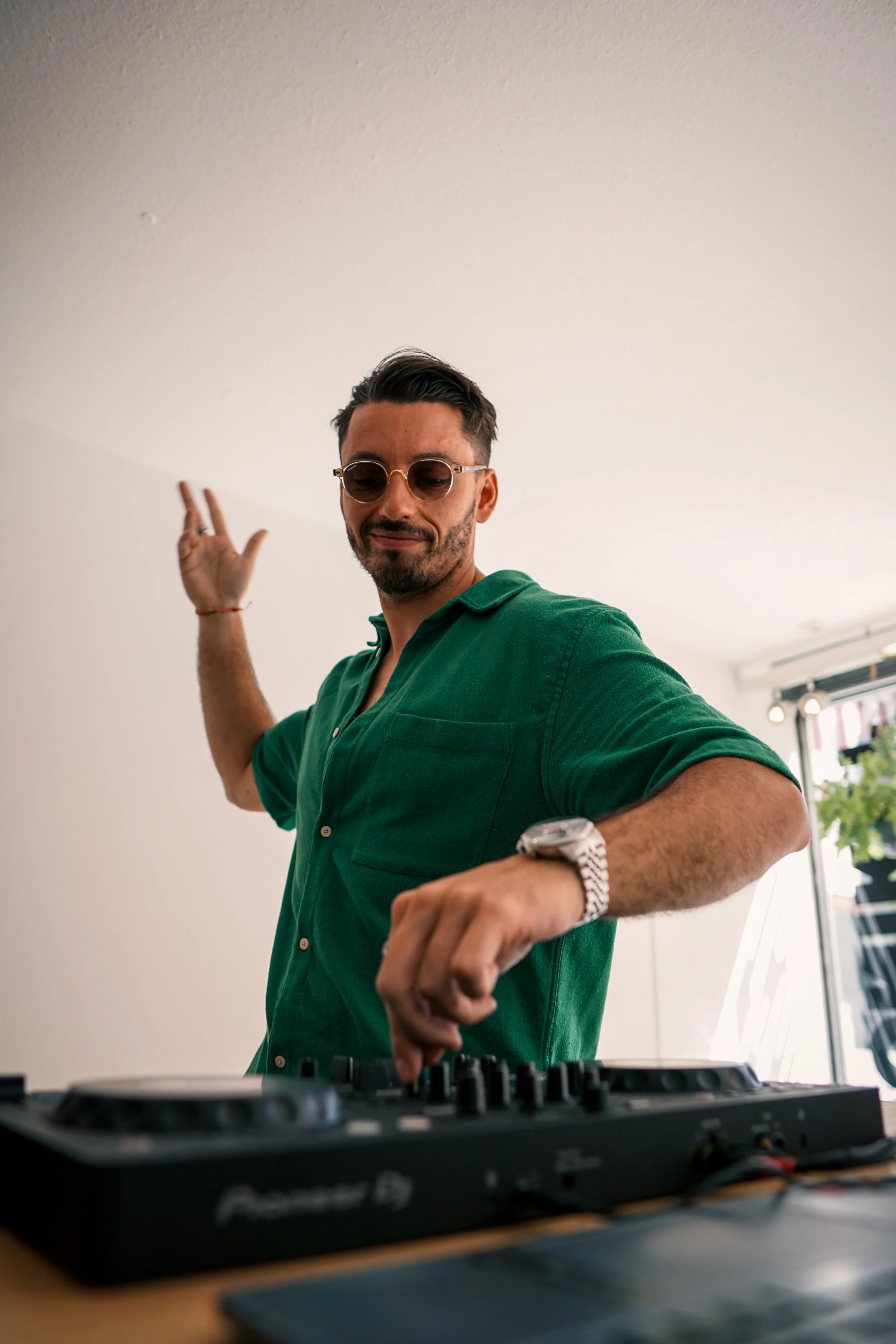 Ein Mann in einer grünen Hemd, trägt Sonnenbrille, ist an einem DJ-Controller und scheint zu tanzen oder zu feiern.