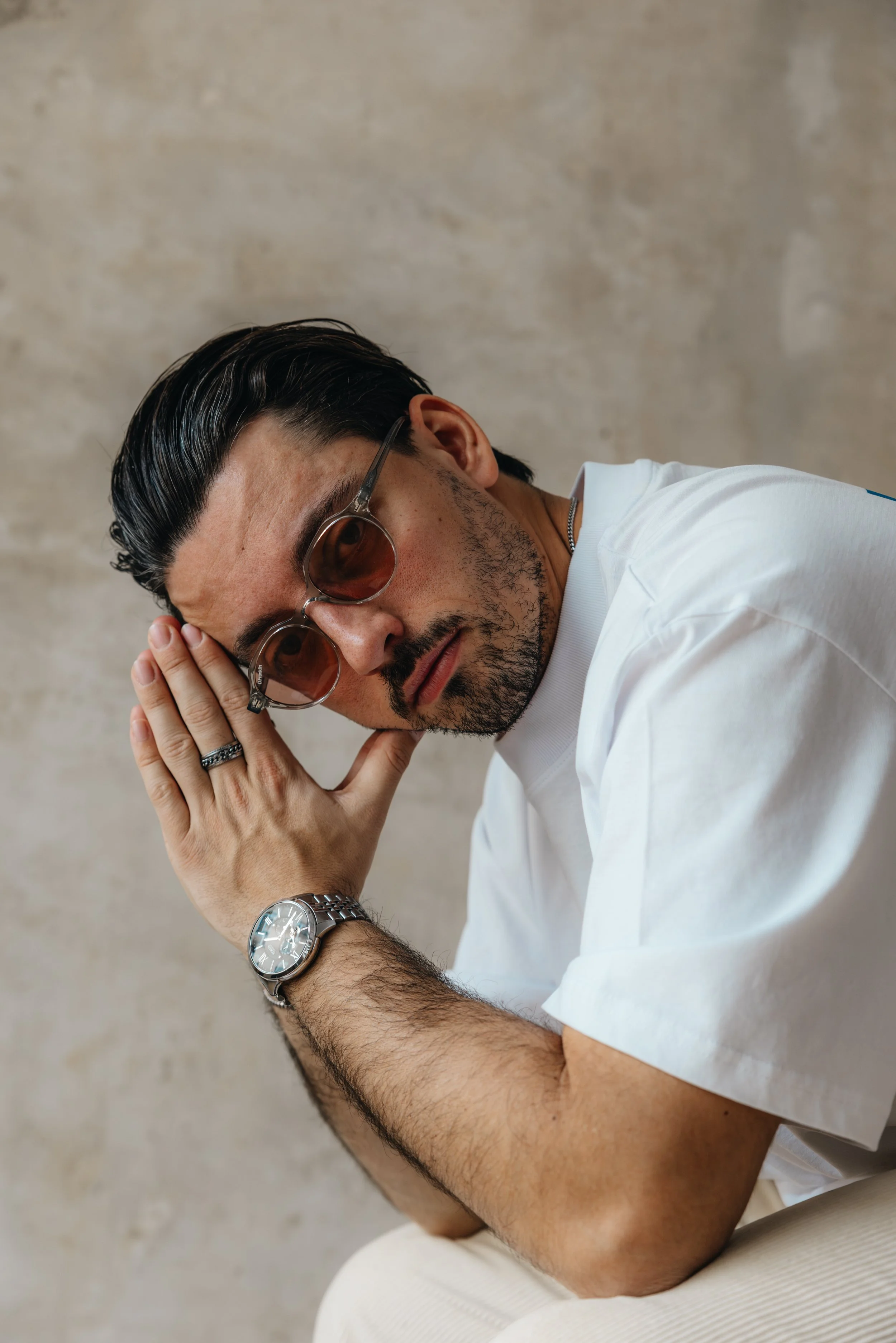 Mann mit dunklen Haaren, Brille und Bart, trägt eine weiße T-Shirt, sitzt gegen eine beige Wand, hält seinen Kopf mit der Hand, trägt eine Armbanduhr, trägt eine Silberkette und einen Ring.