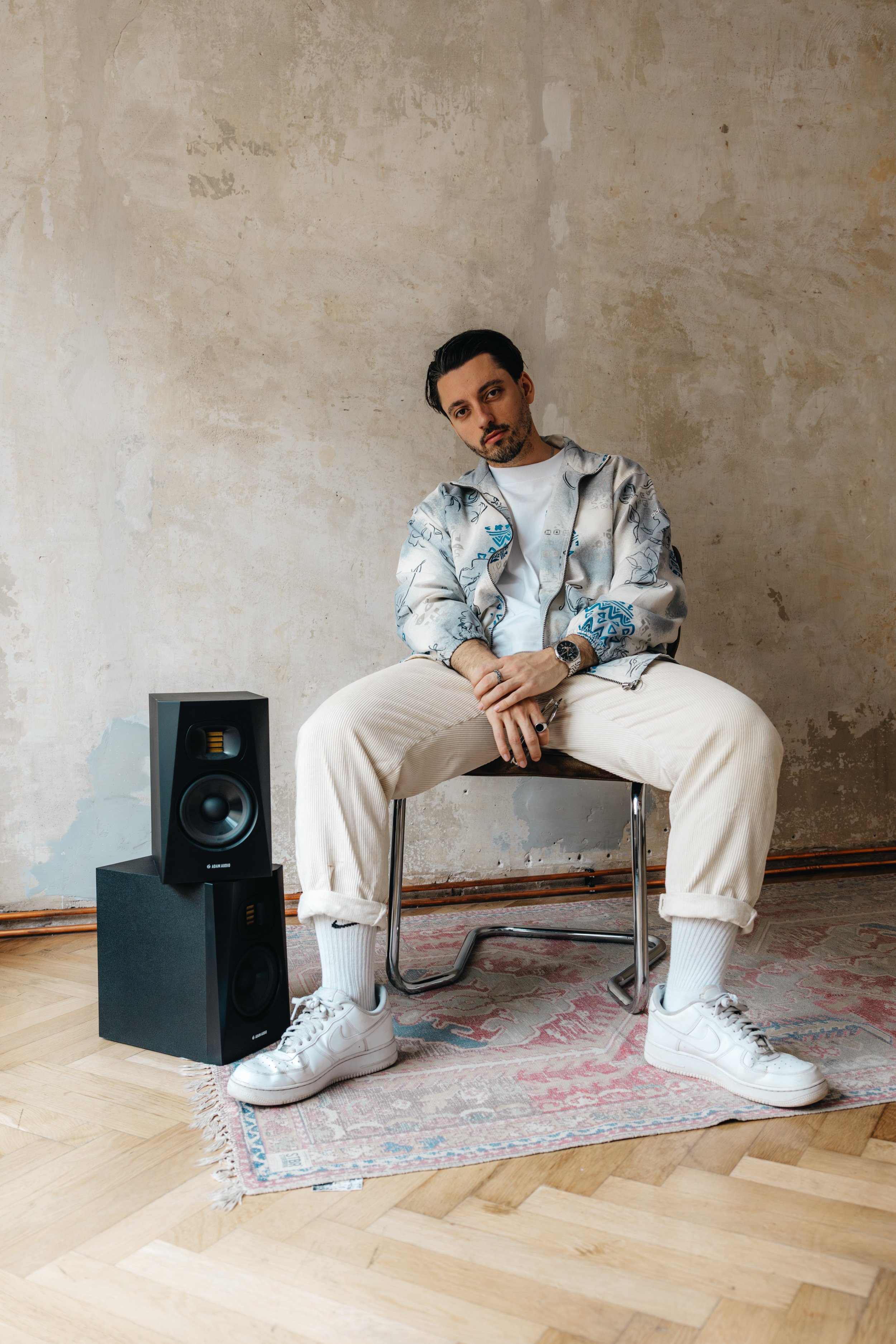 Junger Mann sitzt auf einem Stuhl vor einer alten Wand mit speckigen Verfärbungen, trägt weiße Turnschuhe, beige Cordhosen, weiße Socken, ein weißes T-Shirt und eine silberne Jacke mit blauen Mustern, hält ein Musikinstrument in den Händen, neben ihm