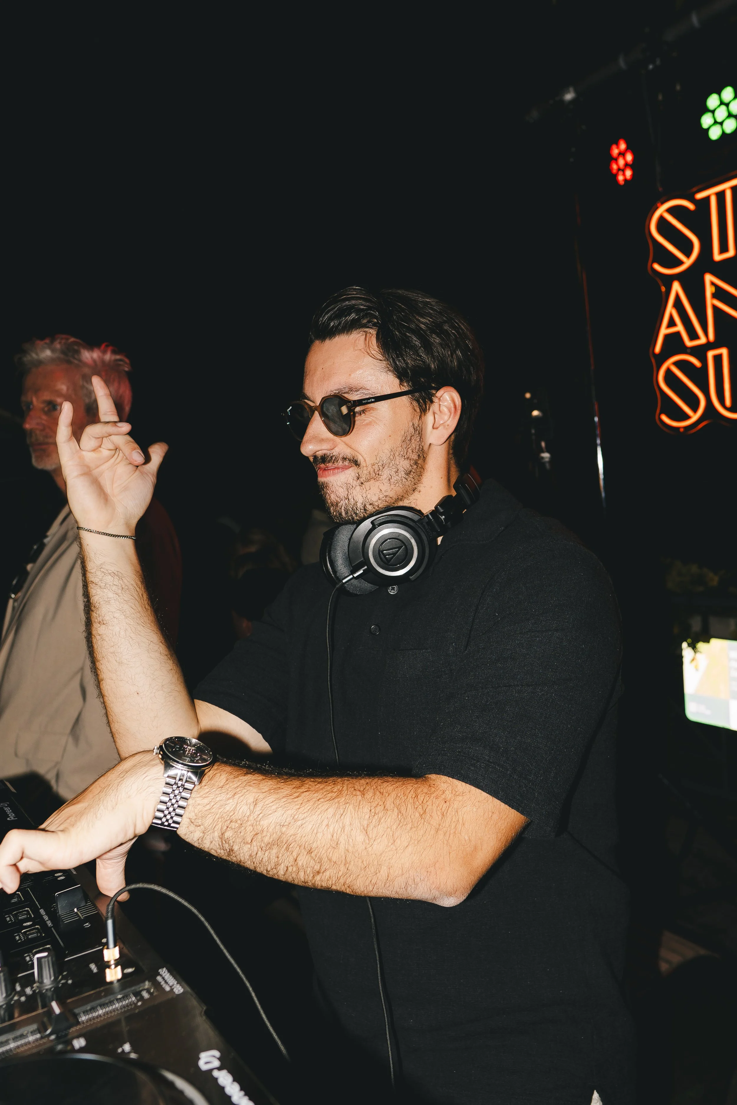 Ein junger Mann mit dunklen Haaren, Sonnenbrille und Kopfhörern, der auf einem DJ-Deck auflegt, während er eine Hand in die Luft hebt. Im Hintergrund sind bunte Lichtstrahler und ein Neon-Schild mit der Aufschrift 'Süß' zu sehen.