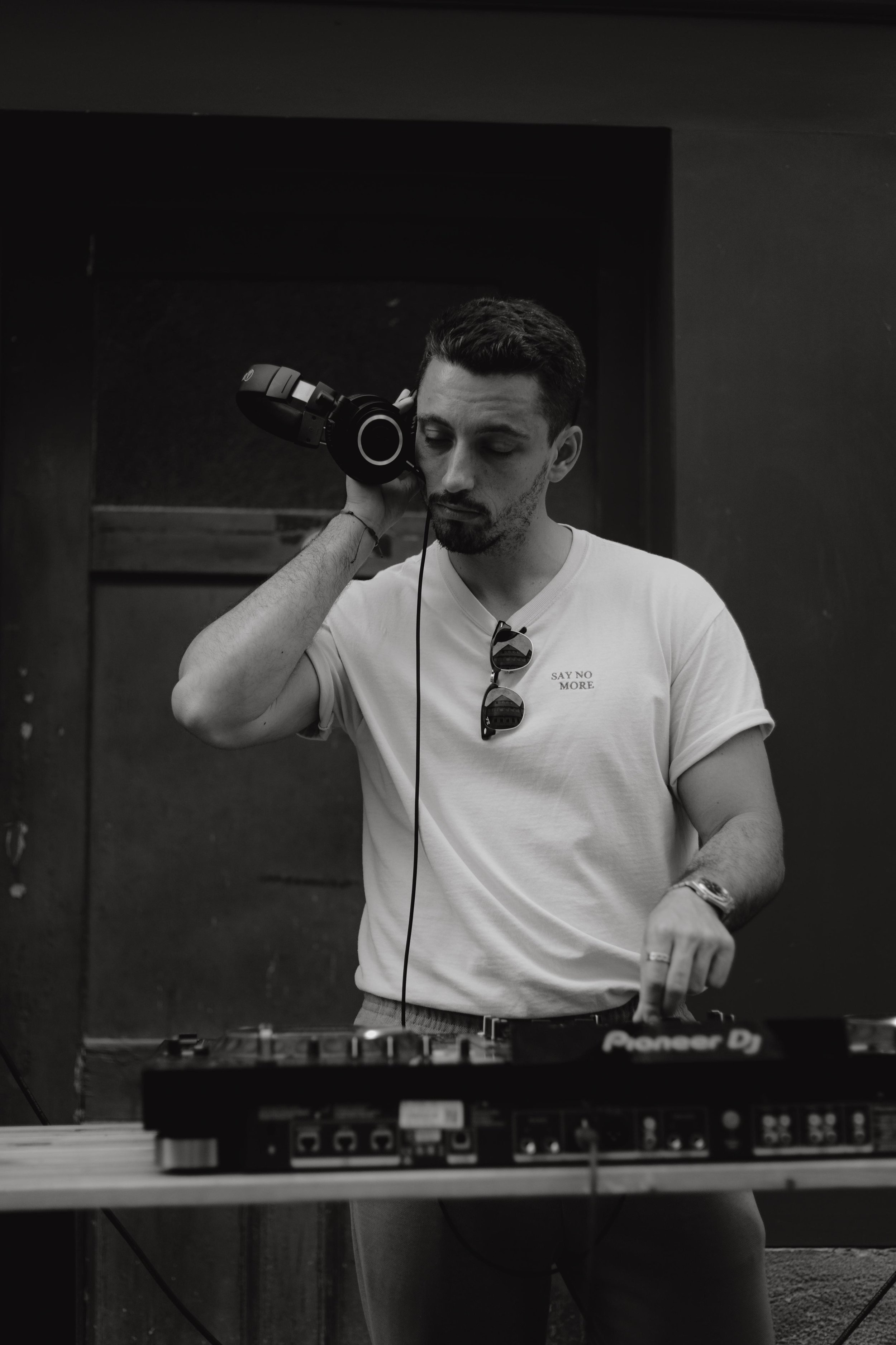 Ein junger Mann mit dunklem Haar und Bart trägt Kopfhörer und steht hinter einem DJ-Controller, mit Sonnenbrille, die an seinem T-Shirt hängt, während er in einem dunklen Raum auflegt.