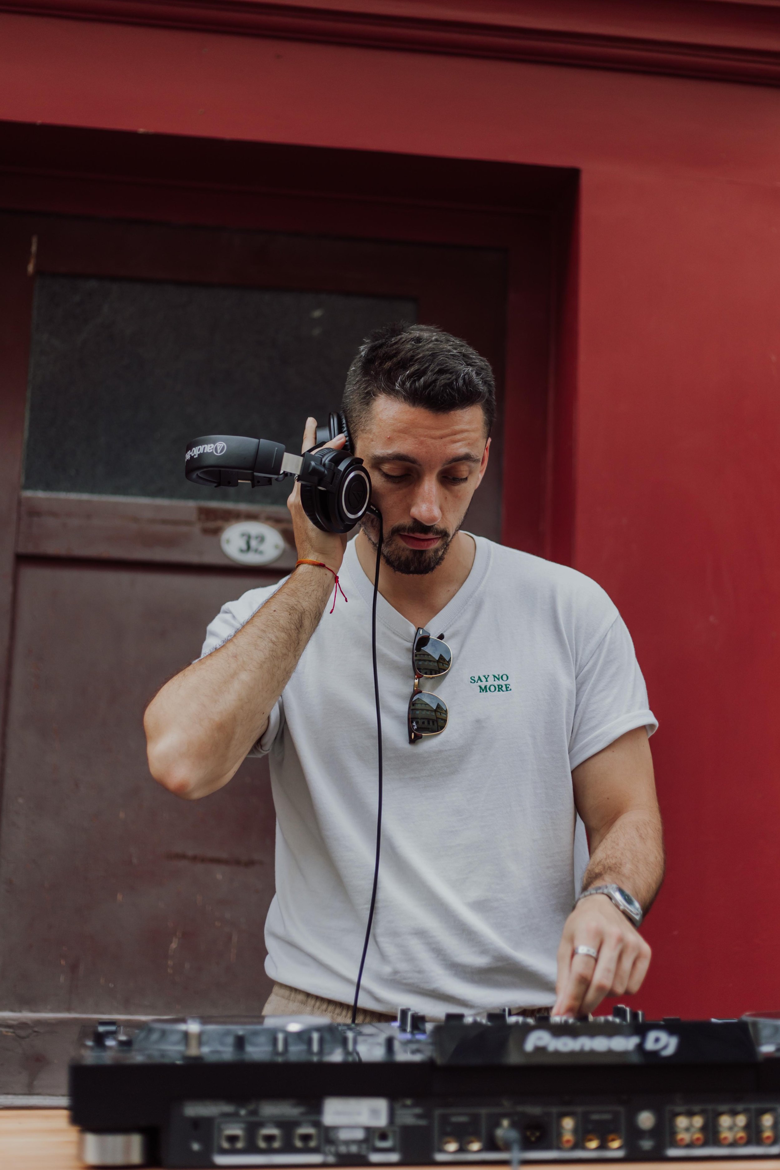 Ein junger Mann mit dunklem Haar und Bart trägt Kopfhörer, während er an einem DJ-Controller arbeitet. Er trägt ein weißes T-Shirt mit grüner Aufschrift 'SAY NO MORE'.