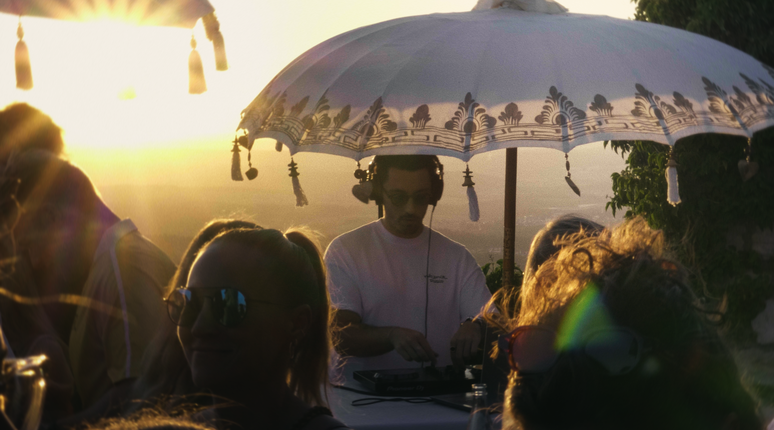 Menschenmenge bei Sonnenuntergang, DJ mit Kopfhörern unter einem weißen Sonnenschirm, der mit Schnörkeln verziert ist, im Freien, umgeben von Bäumen.