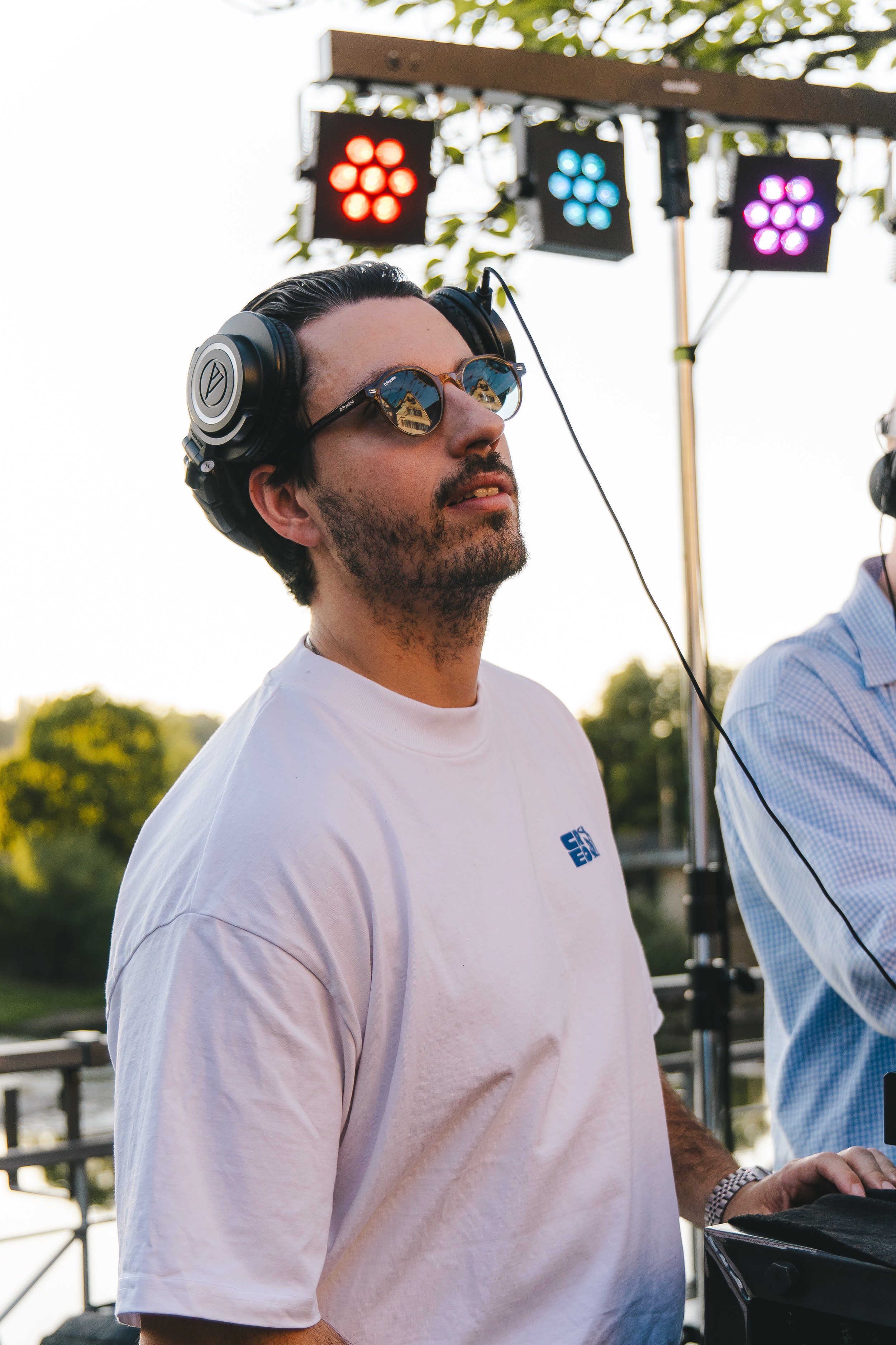 Ein Mann trägt Kopfhörer, Sonnenbrille und ein weißes T-Shirt. Im Hintergrund sind bunte Lichtstrahler. Es scheint eine Outdoor-Veranstaltung bei Sonnenuntergang zu sein.