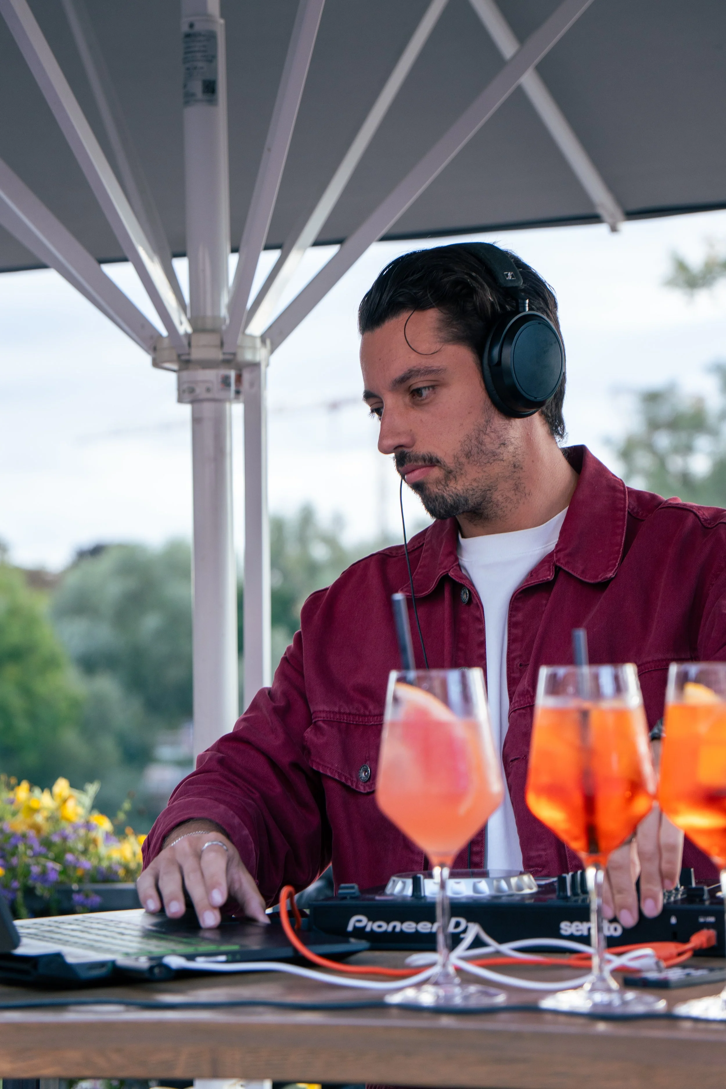Mann mit schwarzen Haaren und Bart, trägt Over-Ear-Kopfhörer, spielt auf einem DJ-Controller an einem Tisch, umgeben von Getränken in Gläsern, im Freien unter einem Pult mit weißen Streben, im Hintergrund Bäume und Himmel.