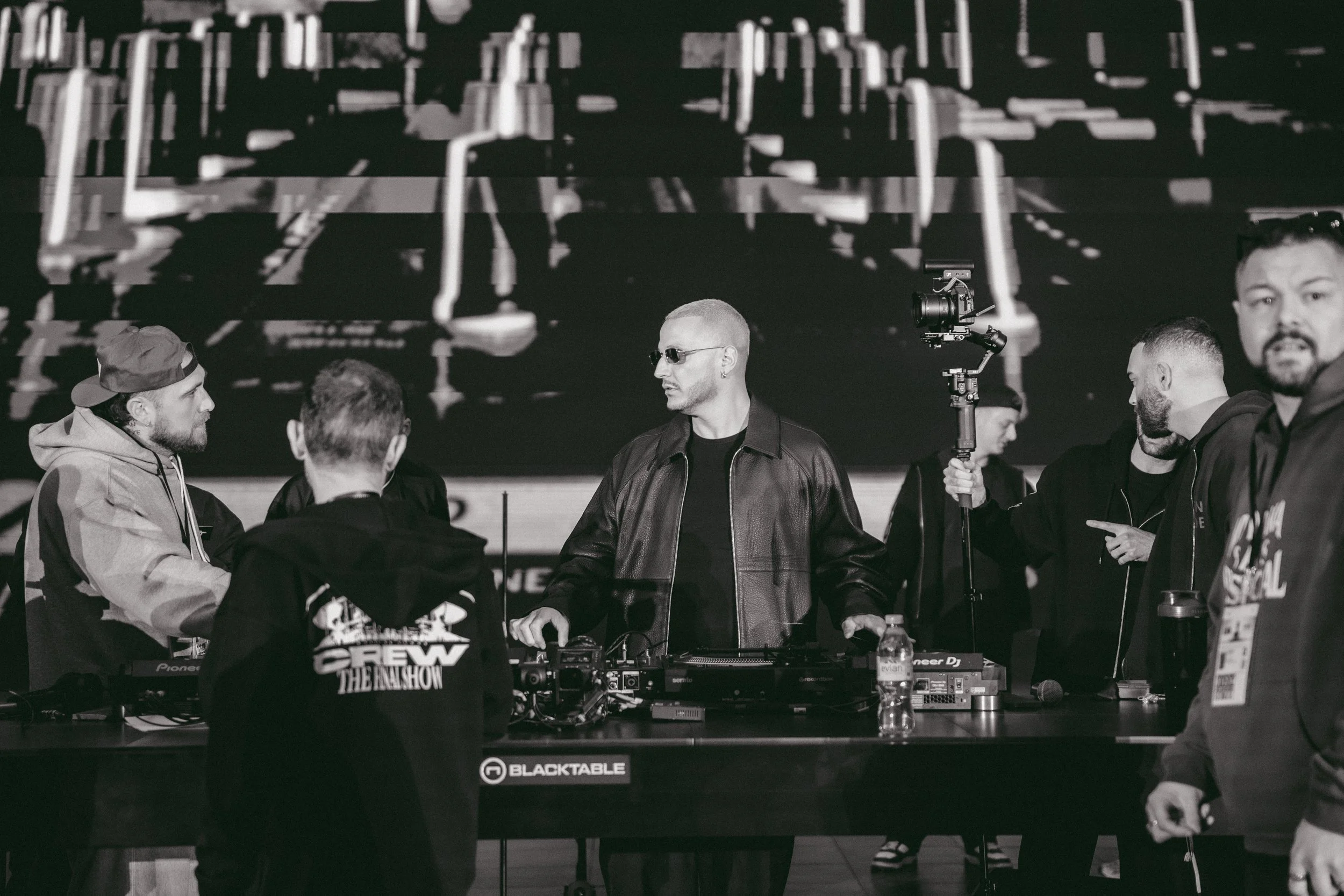 Groupe de personnes dans un stand ou un événement avec un DJ et un écran en arrière-plan, en noir et blanc.
