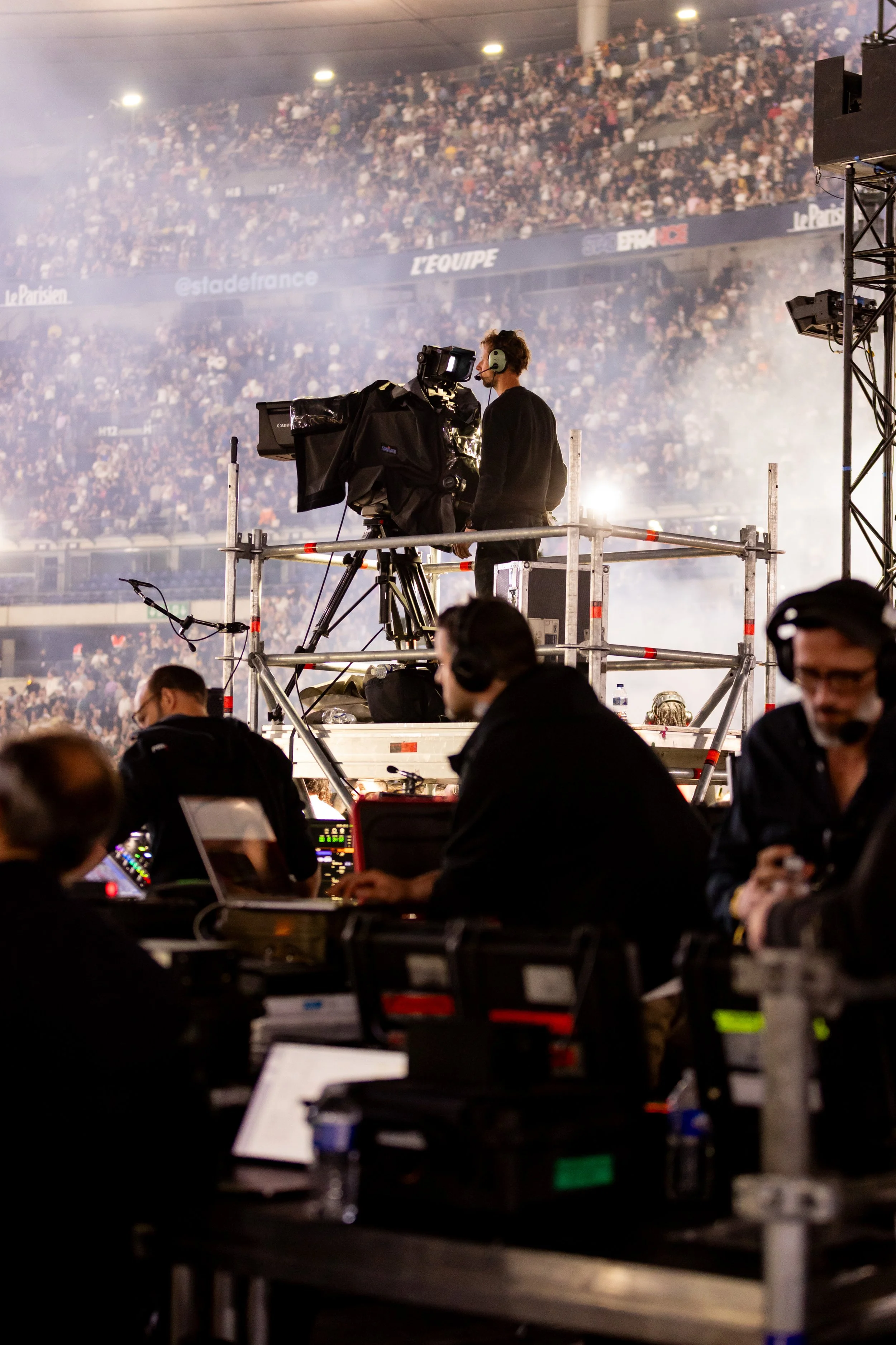Une personne en train de filmer un événement sportif dans un stade rempli de spectateurs, avec du matériel de tournage sur une plateforme élevée, et une grande foule dans les tribunes.