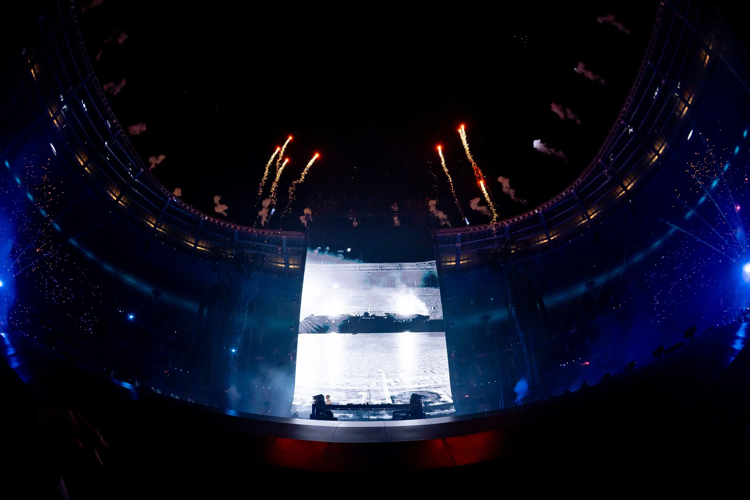 Un concert ou fête dans une grande salle, avec des feux d'artifice dans le ciel noir.