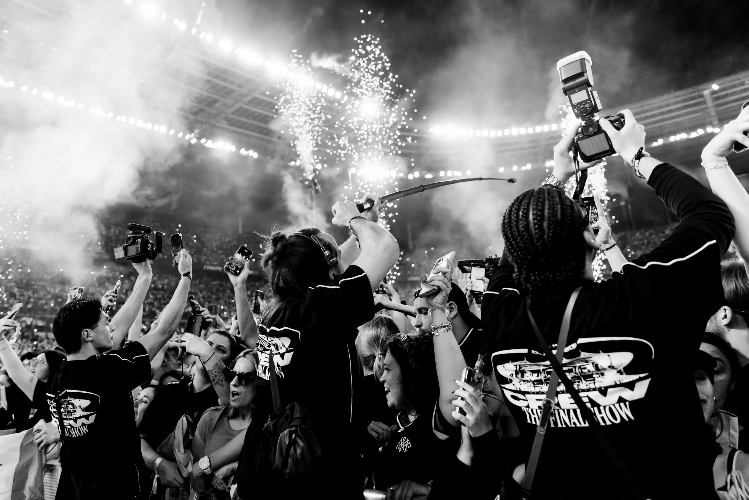 Une scène de concert ou d'événement avec un grand groupe de personnes, beaucoup de femmes, qui prennent des photos ou filment avec leur téléphone. En arrière-plan, des feux d'artifice ou des effets lumineux dans un stade ou une arène.