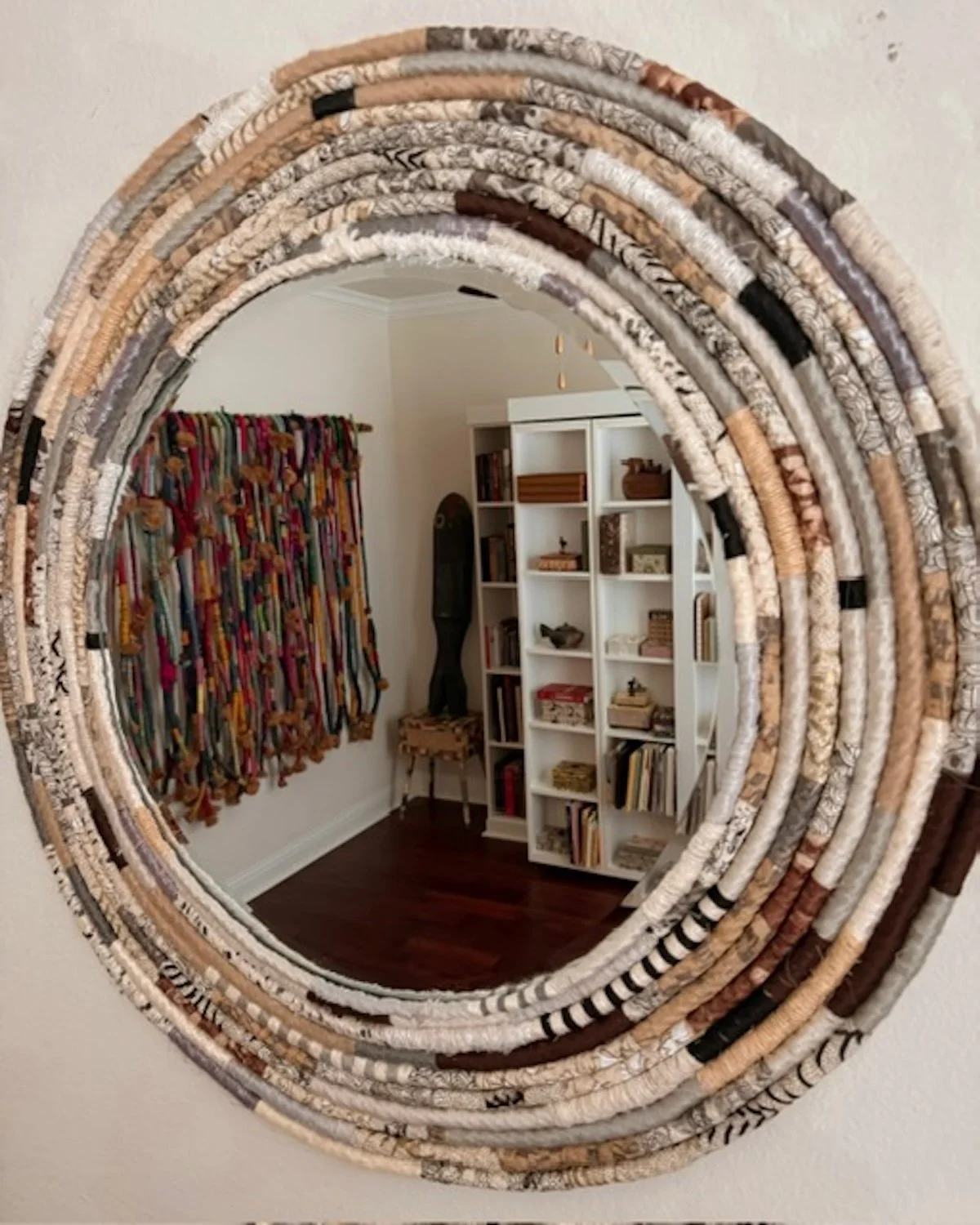 A round mirror with a frame made of rolled fabric strips reflects a cozy room with a white bookshelf, a dark wooden floor, a colorful decorative curtain, and a small wooden stool.