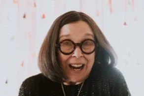 Woman with shoulder-length dark hair, wearing large round glasses and a black top, smiling excitedly in front of a white curtain with red and orange decorations.