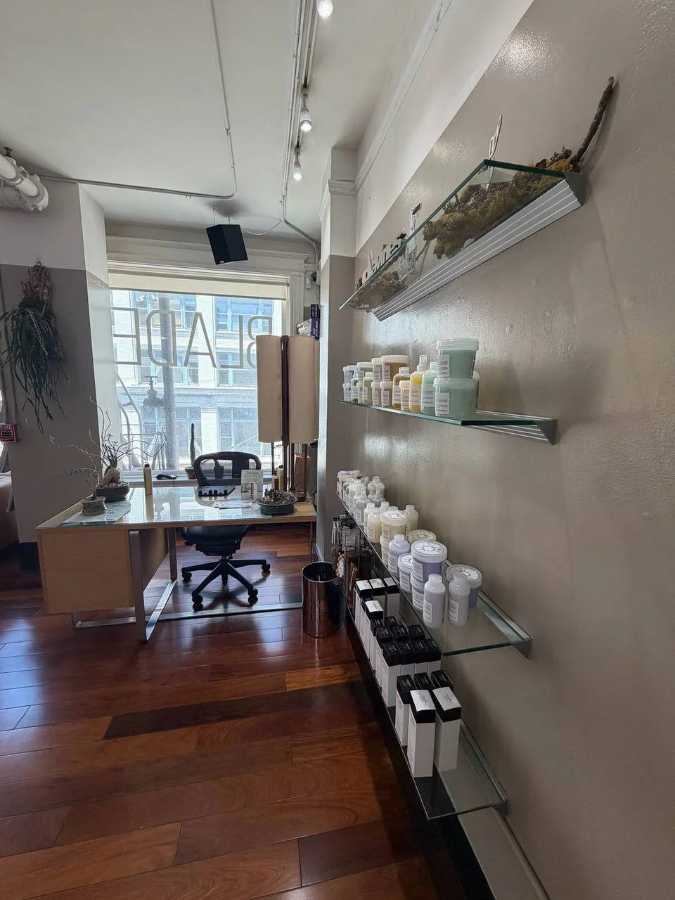 Interior of a wellness or treatment room with a desk and an office chair near a window. Shelves on the wall hold various bottles and containers of supplements or skincare products. The room has wooden flooring and natural light streaming in through the window.