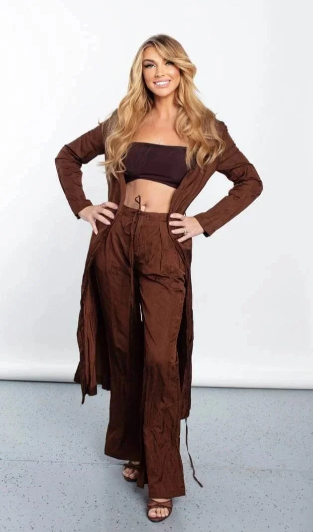 Woman with long blonde hair smiling, wearing a black crop top, brown jacket, and matching wide-leg pants, standing against a white background.
