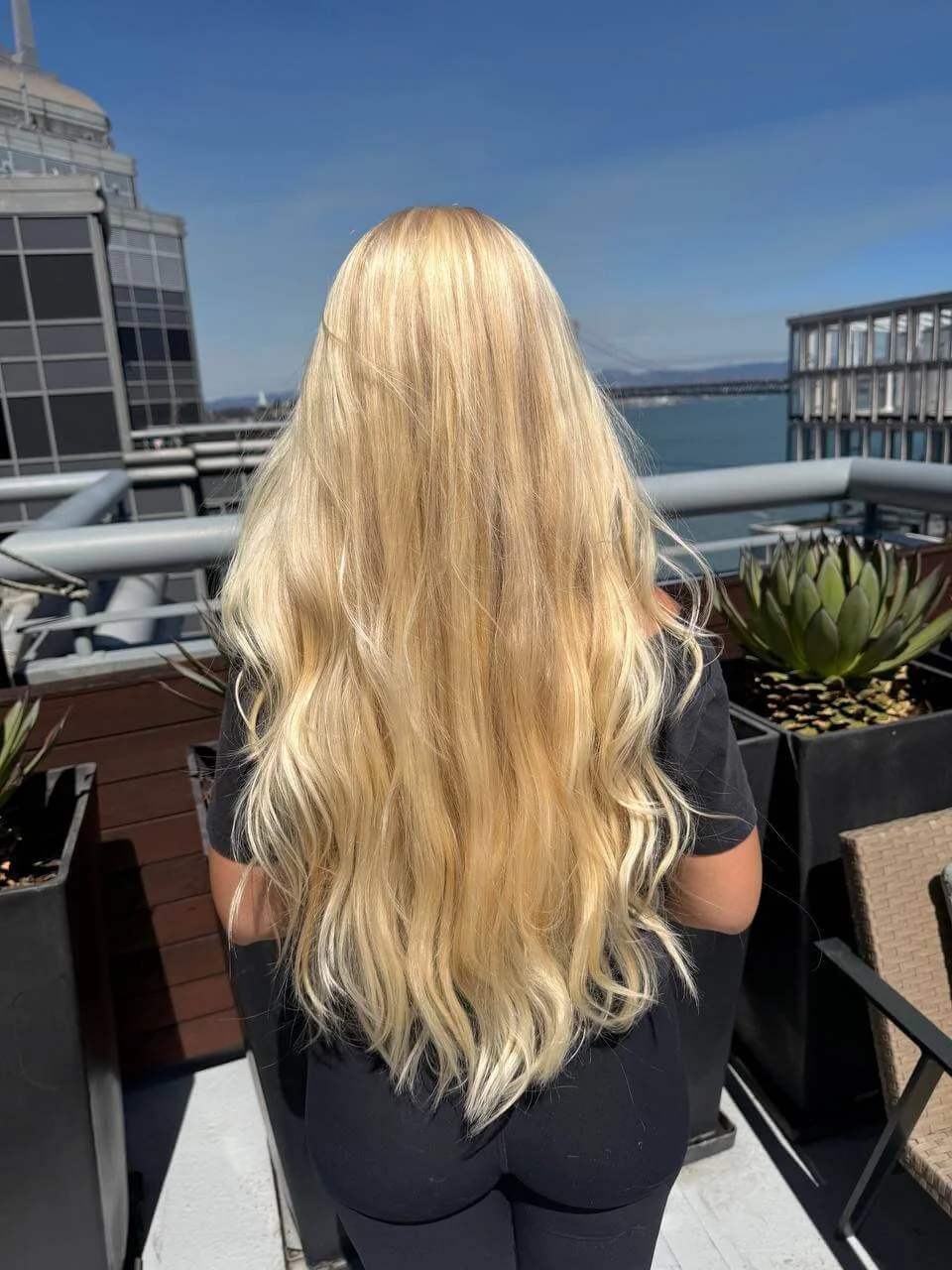 Back view of a woman with long, blonde wavy hair standing on a balcony with a view of a city skyline and water.