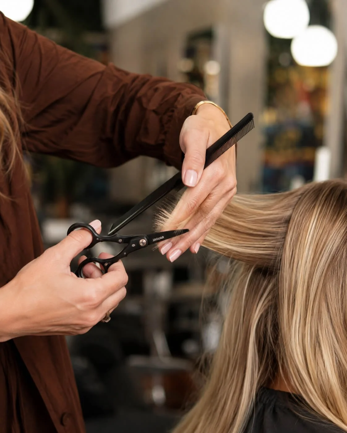 A hairstylist cutting a woman's blonde hair with scissors while holding the hair with a comb.