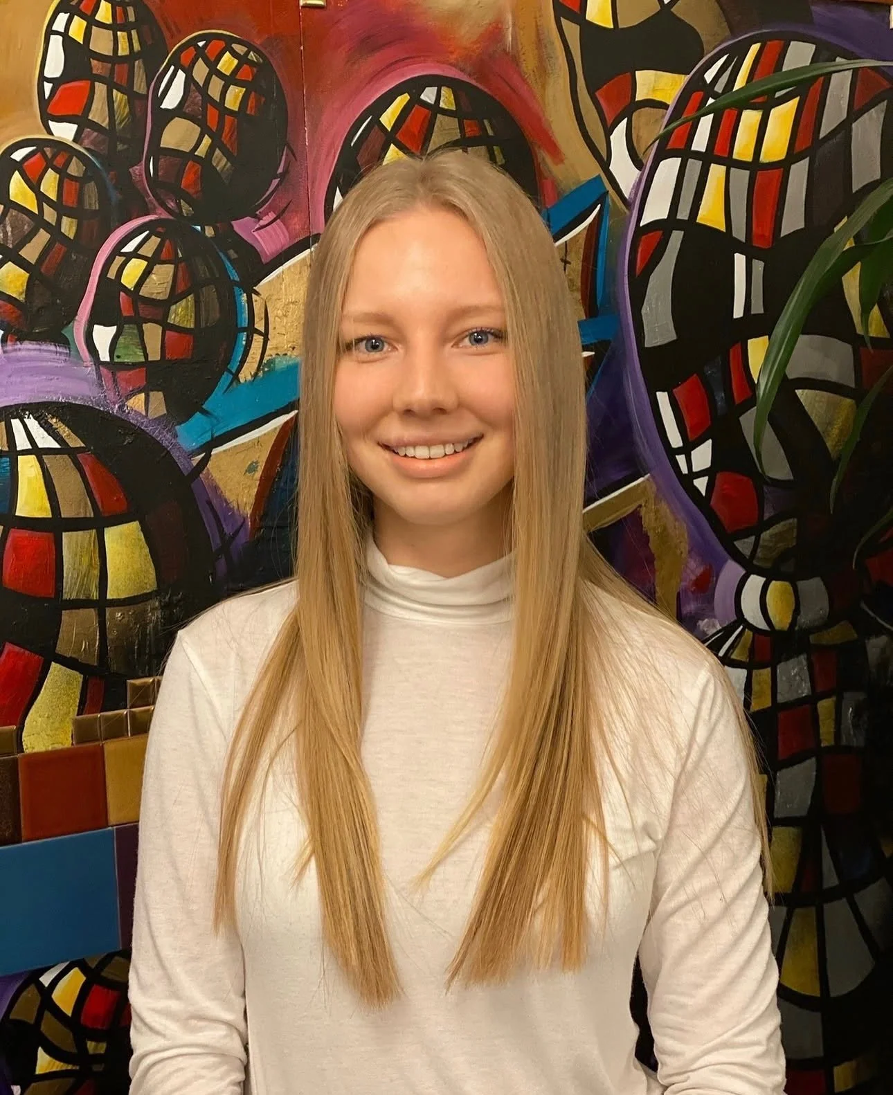 A young woman with long blonde hair and blue eyes smiling, standing in front of a colorful abstract mural with mosaic-like patterns.