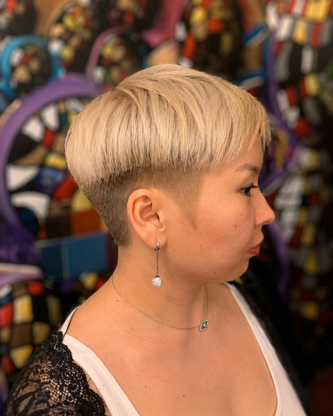Side profile of a woman with a short, platinum blonde hairstyle standing in front of a colorful, abstract graffiti wall.