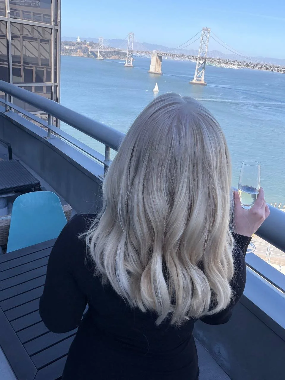 A woman with blonde wavy hair holding a glass of wine on a balcony overlooking a waterway with a bridge in the distance.