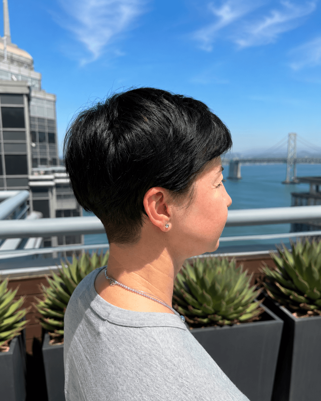 A woman with short black hair and earrings standing on a balcony overlooking a river and a bridge under a clear blue sky.