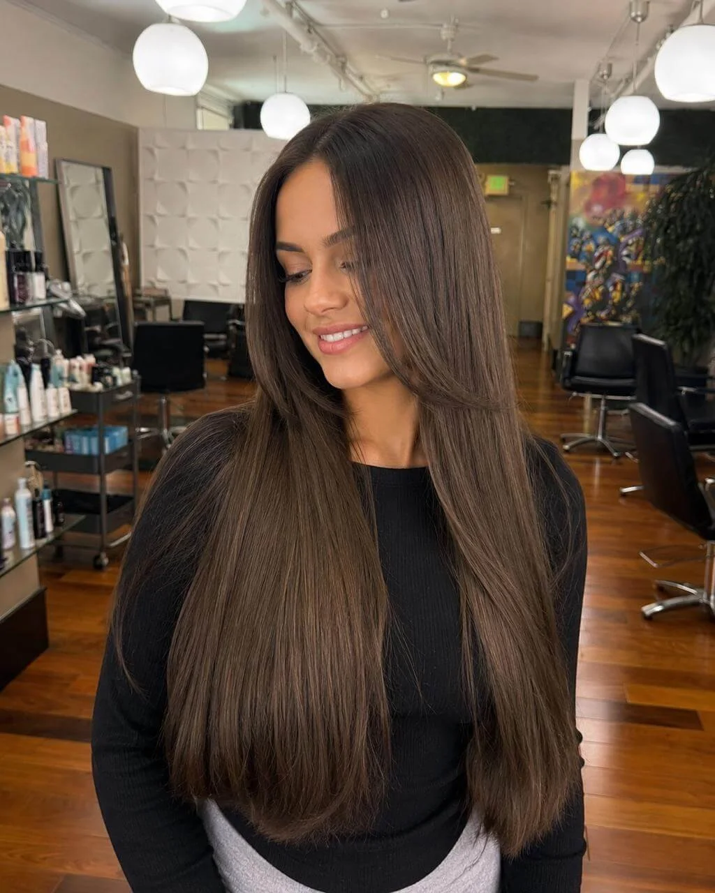 A young woman with long, straight brown hair and light makeup inside a hair salon, smiling softly with eyes closed.