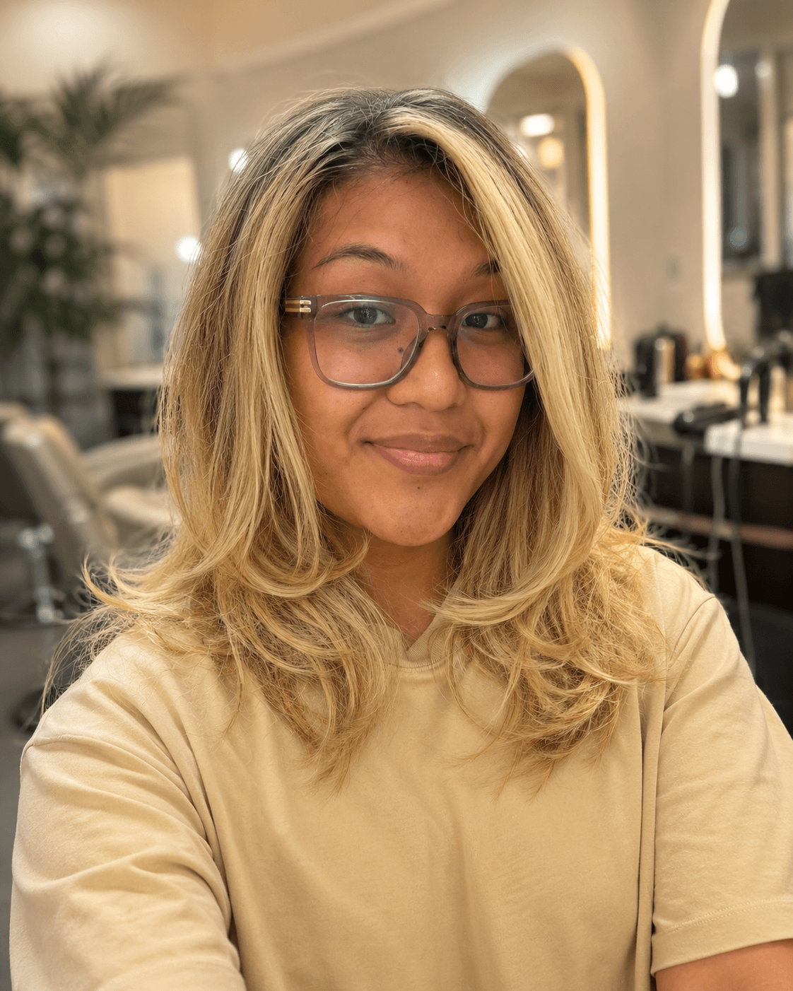 A young woman with blonde, wavy hair and glasses smiling at the camera in a salon or beauty parlor with mirrors and salon equipment in the background.