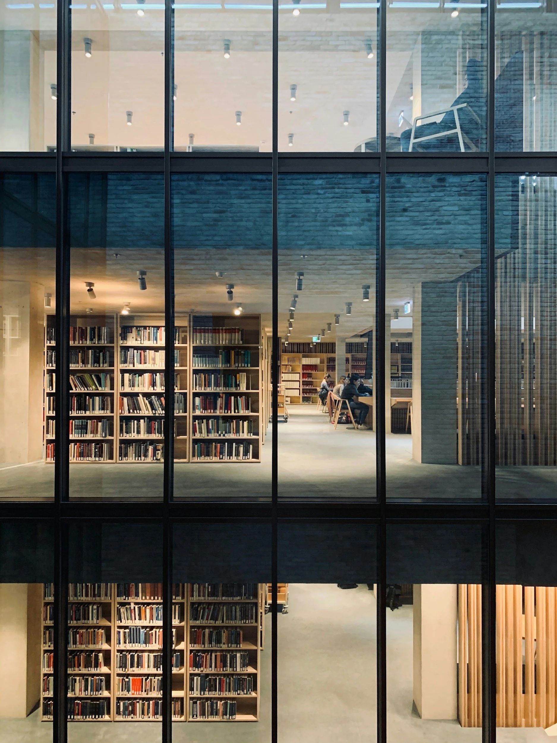 A view through large glass windows showing an interior library with bookshelves, people reading at tables, and a modern architectural design with wood and brick elements.