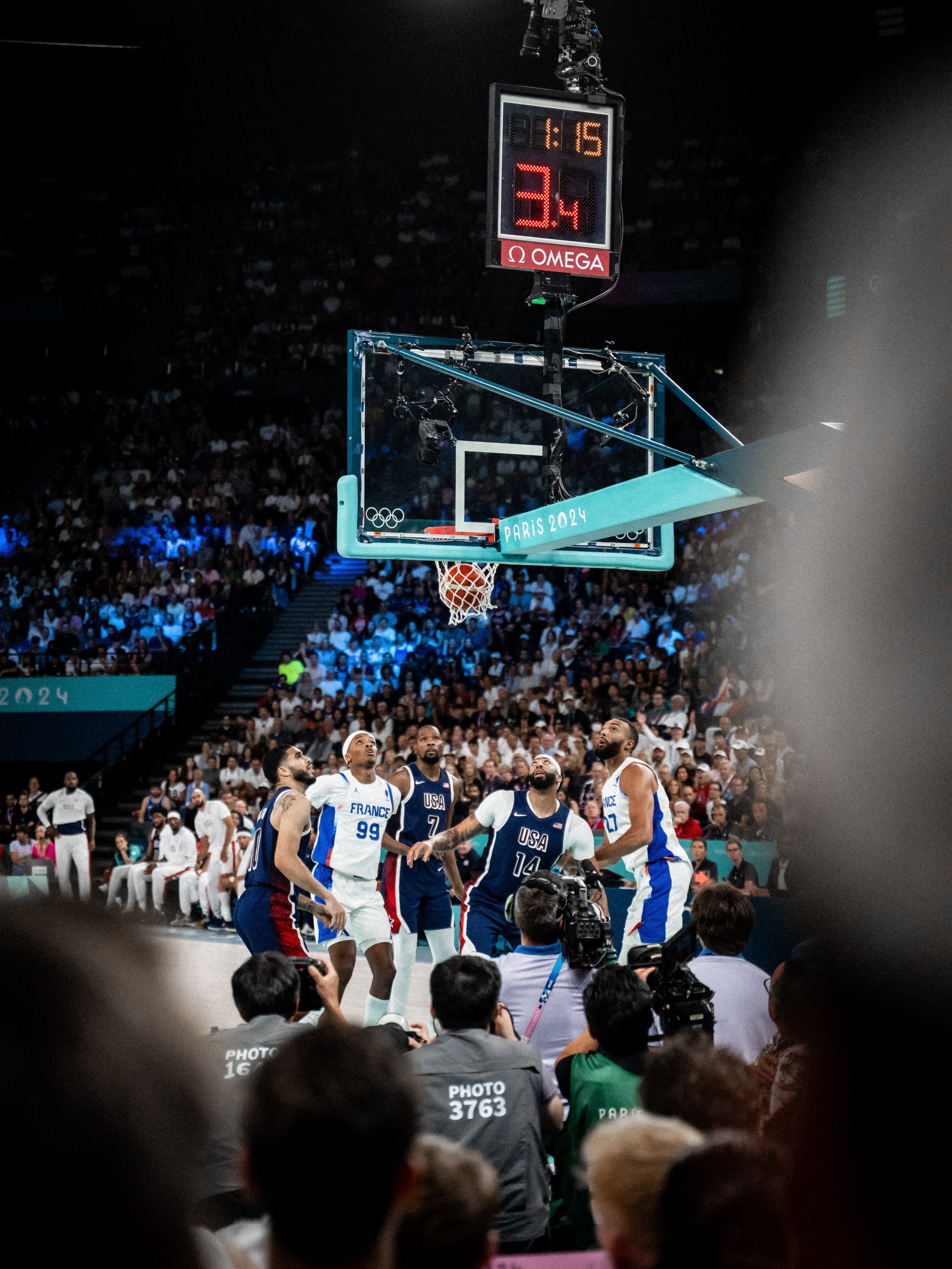 Une scène de basket-ball lors des Jeux Olympiques de Paris 2024. Des joueurs de l'équipe de France et des États-Unis se tiennent près du panier, sous un tableau de scoreboard affichant 8:15 et 3.4 secondes. La foule et plusieurs photographes entourent la scène.