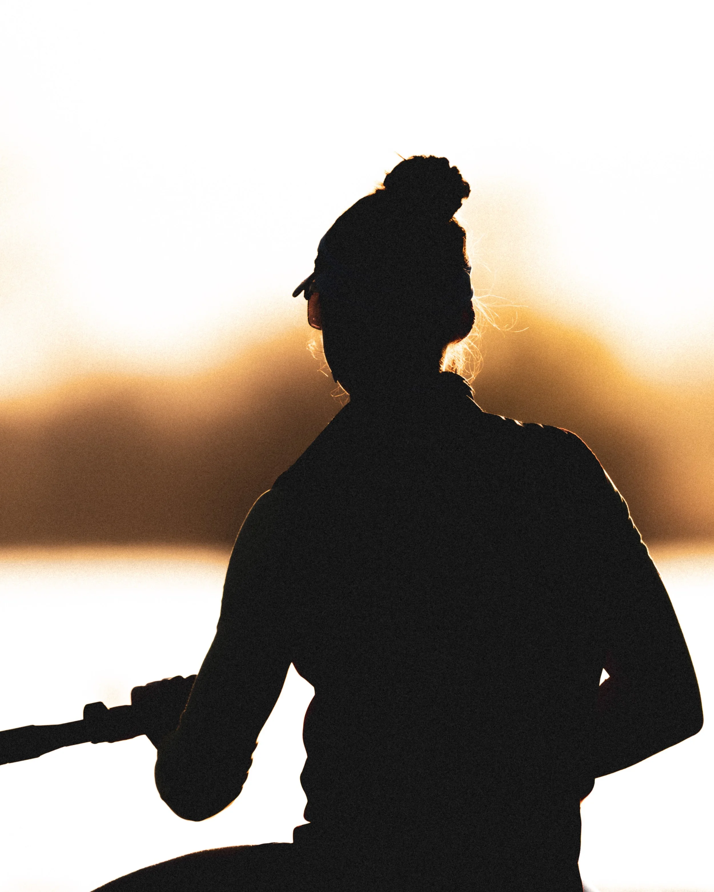 Silhouette d'une personne avec des cheveux bouclés, portant un manteau, lors du coucher de soleil, avec un fond clair et chaud.