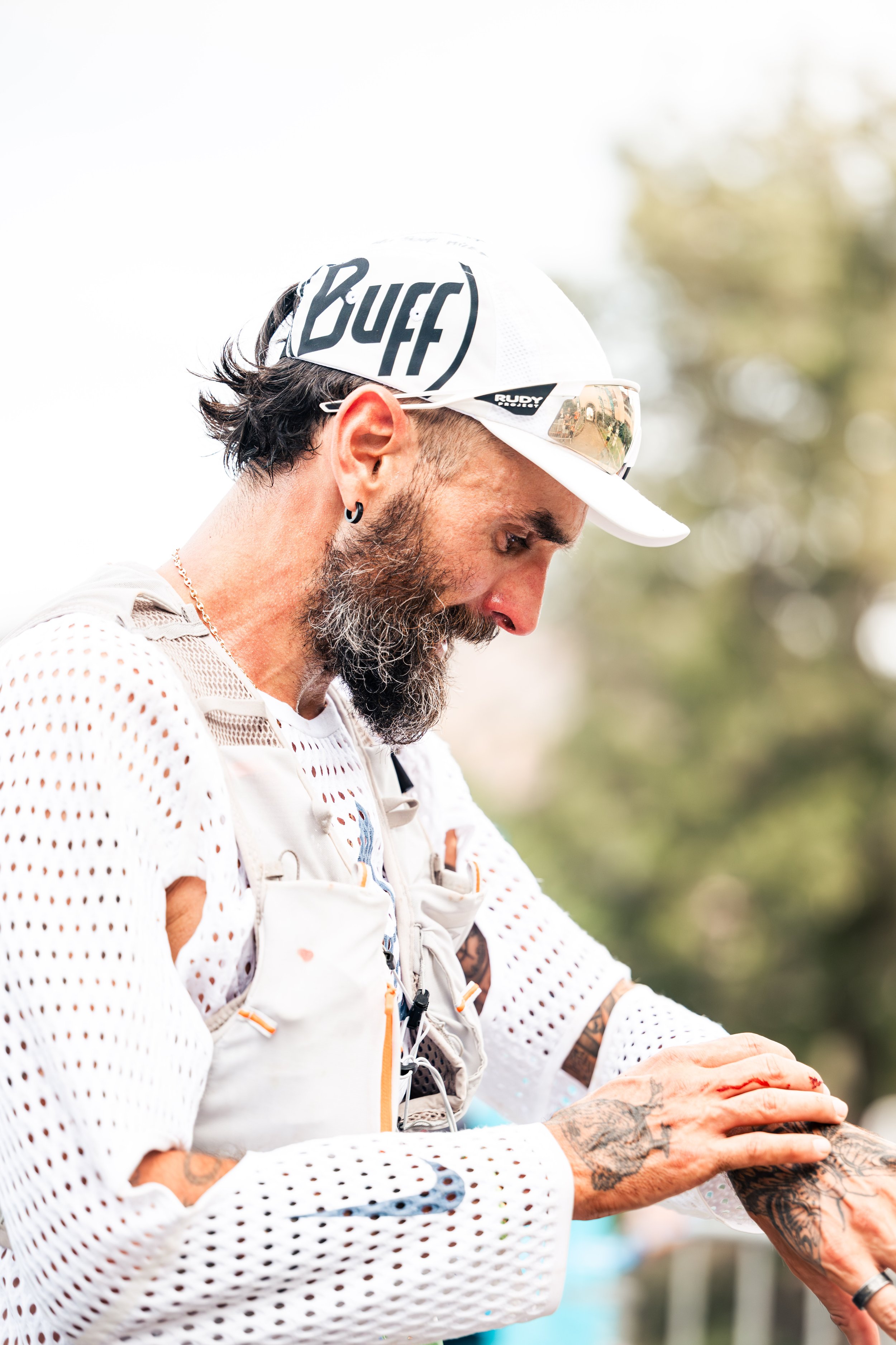 Un homme avec une barbe, portant une casquette blanche avec le mot "Buff" et des lunettes de soleil, prépare une injection ou un vaccin, ses mains tatouées montrent une pointe d'injection près du bras.