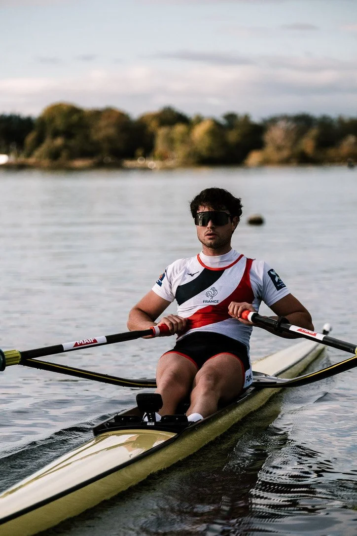 Un homme en tenue de sport d'aviron, portant des lunettes de soleil noires, ramant sur un plan d'eau avec des arbres en arrière-plan.