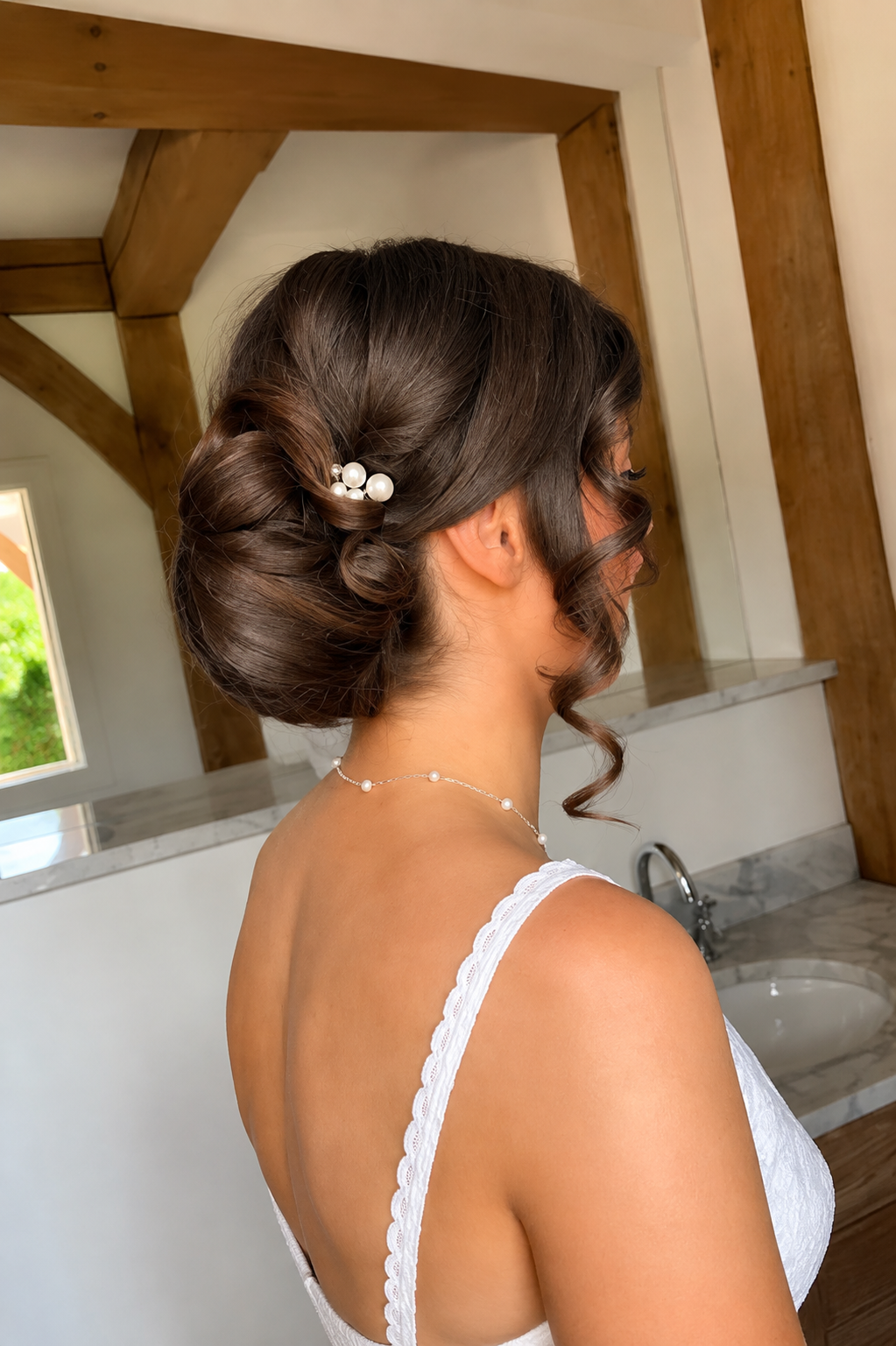 A woman with dark brown hair styled in an elegant updo, decorated with white pearl hairpins, wearing a white lace dress and a pearl necklace, standing in a room with wooden beams and a marble countertop.
