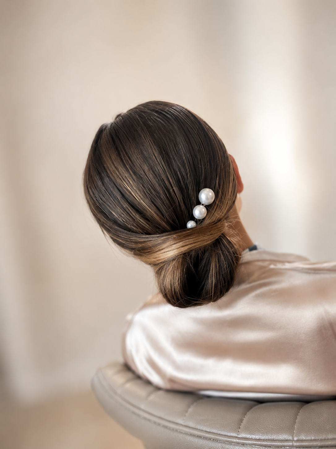 Close-up of a woman's elegant hairstyle with brown hair styled in a smooth bun, decorated with white pearl hairpins, wearing a satin beige garment, sitting on a padded chair against a neutral background.
