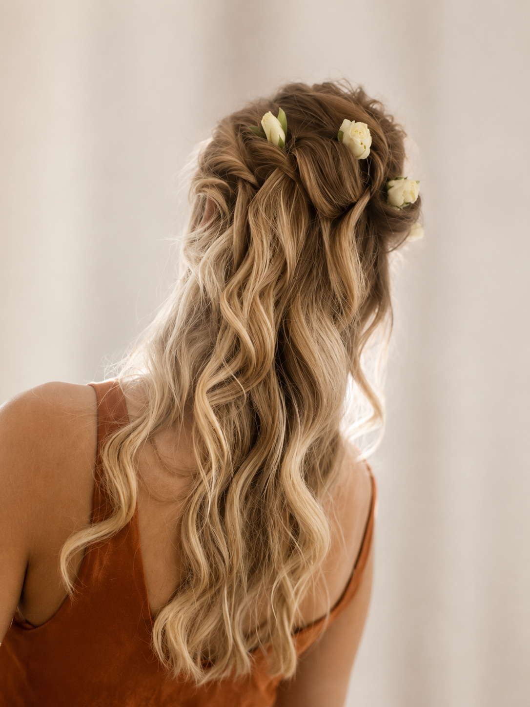 A woman with long, curly blonde hair decorated with white flowers, wearing a sleeveless brown top, facing away from the camera.
